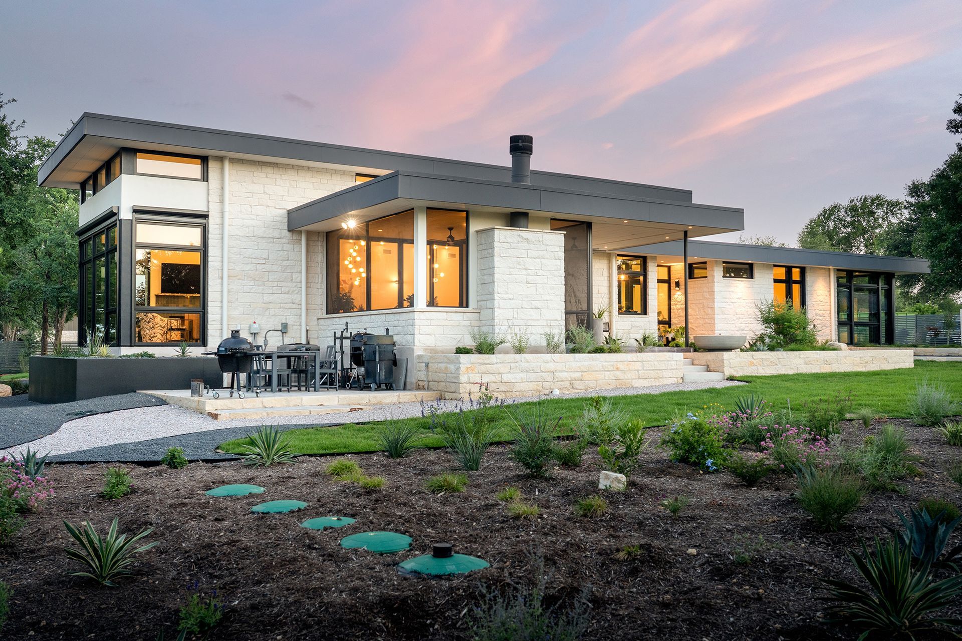 Modern, white brick house with dark trim and large windows, surrounded by a landscaped yard at dusk.
