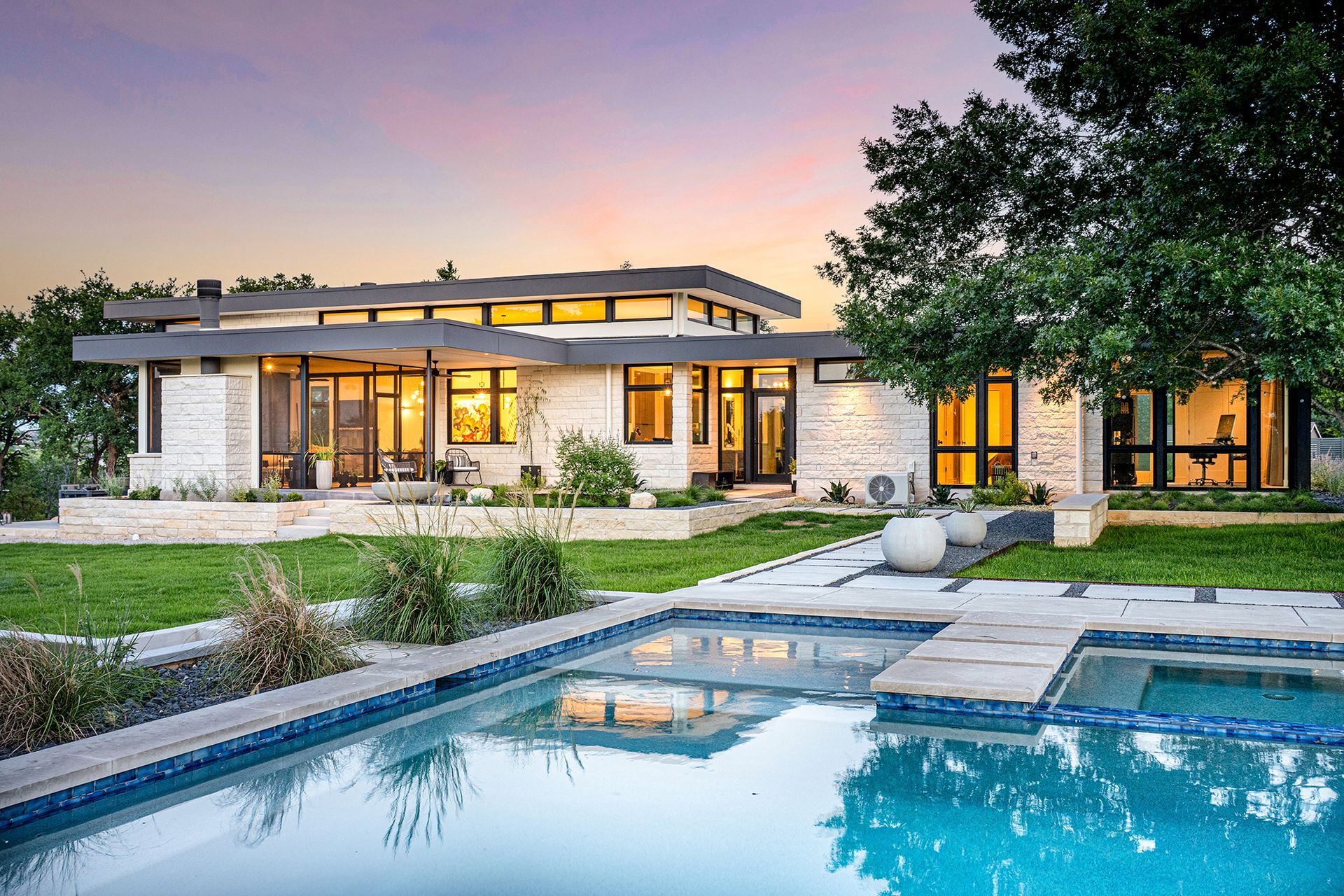 Modern home with a pool at dusk; stone facade, large windows, green lawn, and trees.