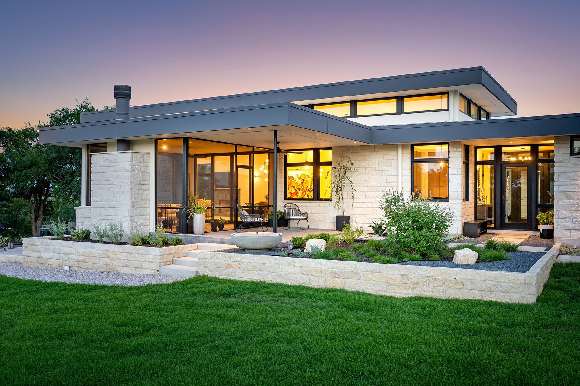 Modern home exterior with light-colored stone walls, dark trim, and a flat roof; features a patio and landscaped yard.