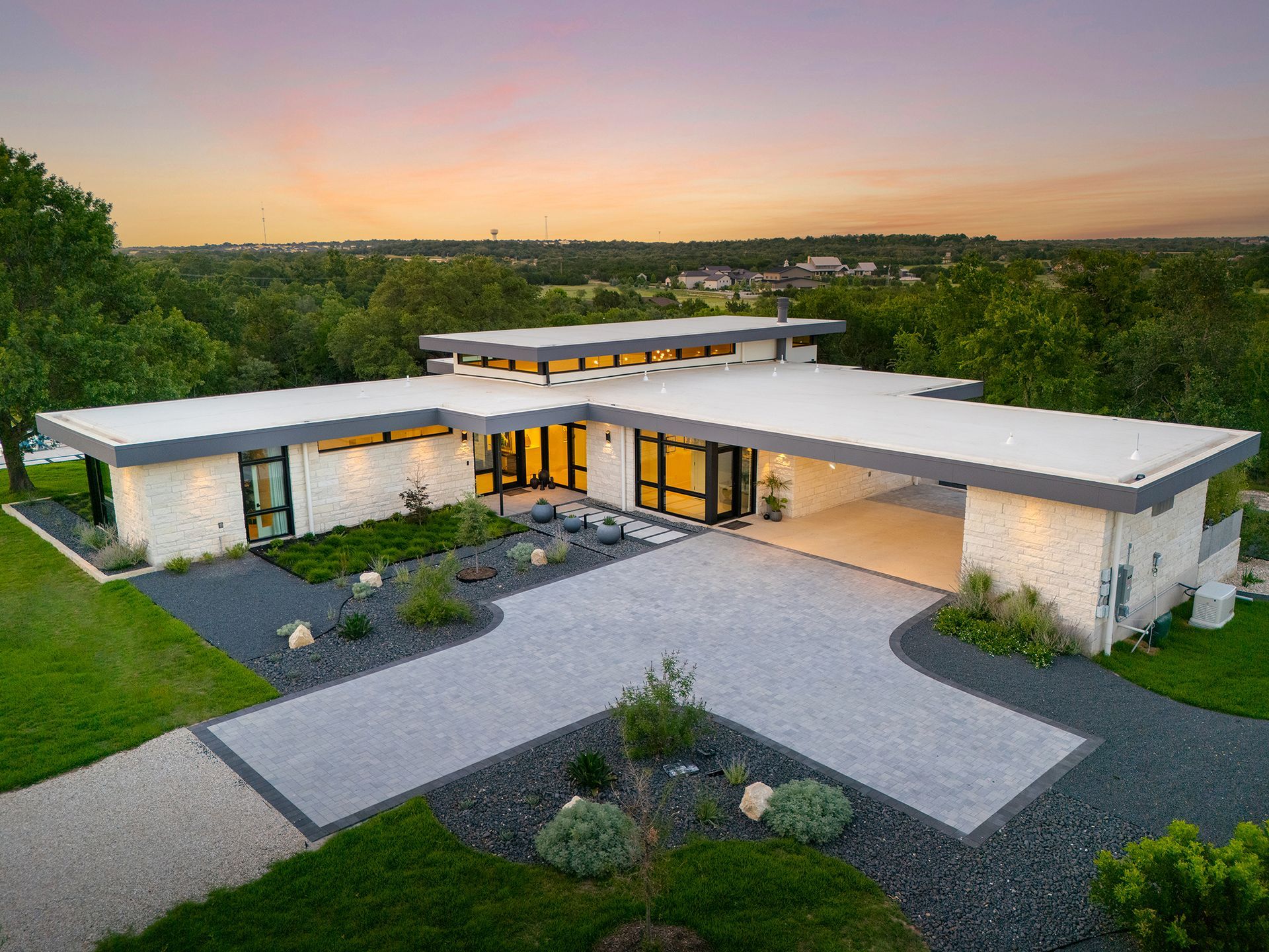 Modern, white-stone house with a flat roof, carport, and gray driveway. Surrounded by landscaping and a scenic view.