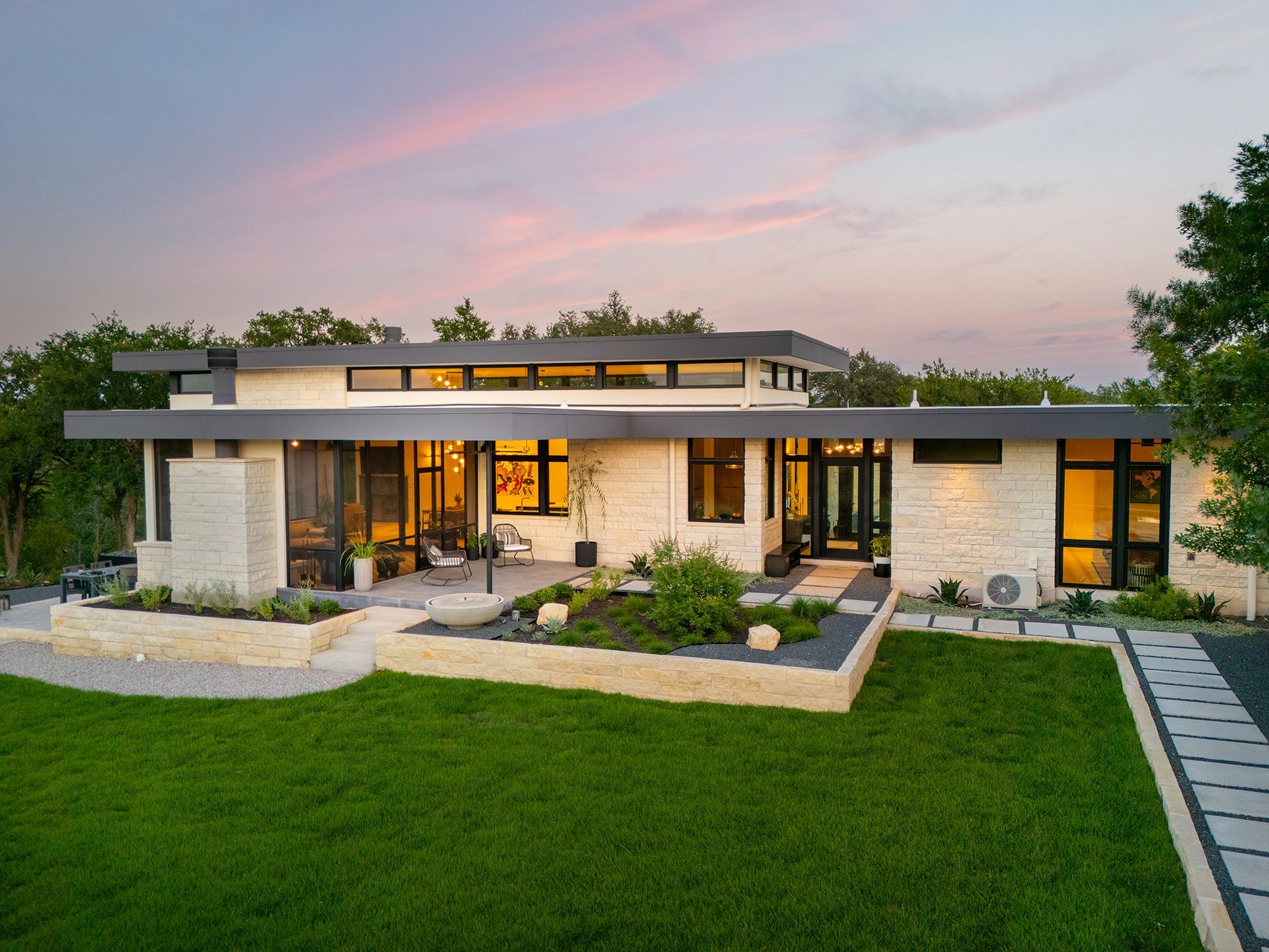 Modern home with stone facade, dark trim, and flat roof at dusk with pink sky.