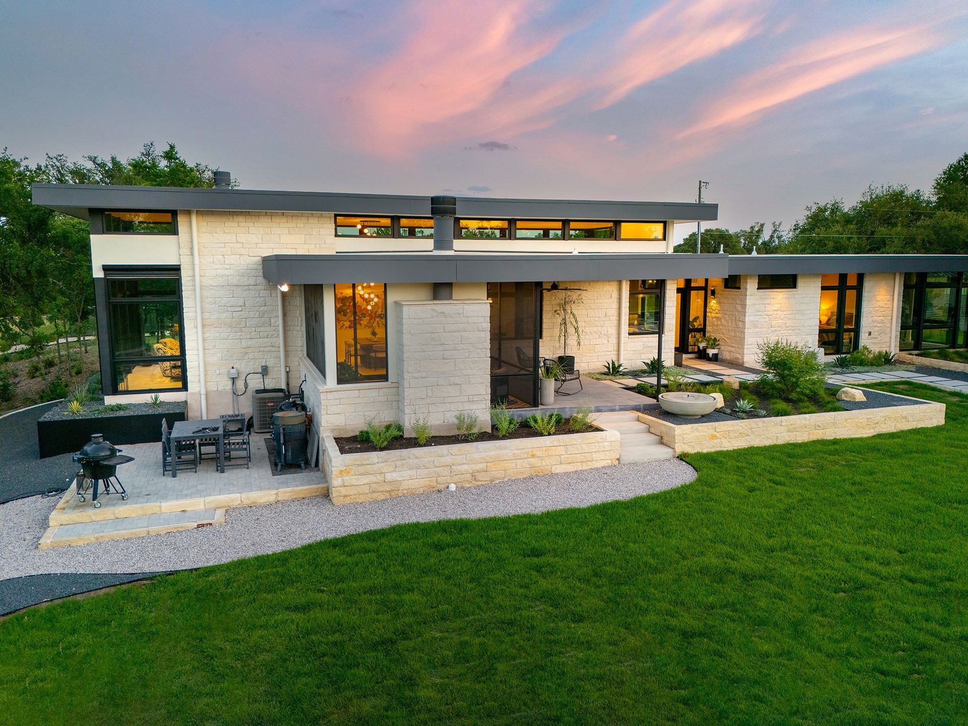 Modern home exterior at sunset. Light-colored stone walls, dark trim, large windows, and manicured landscaping.