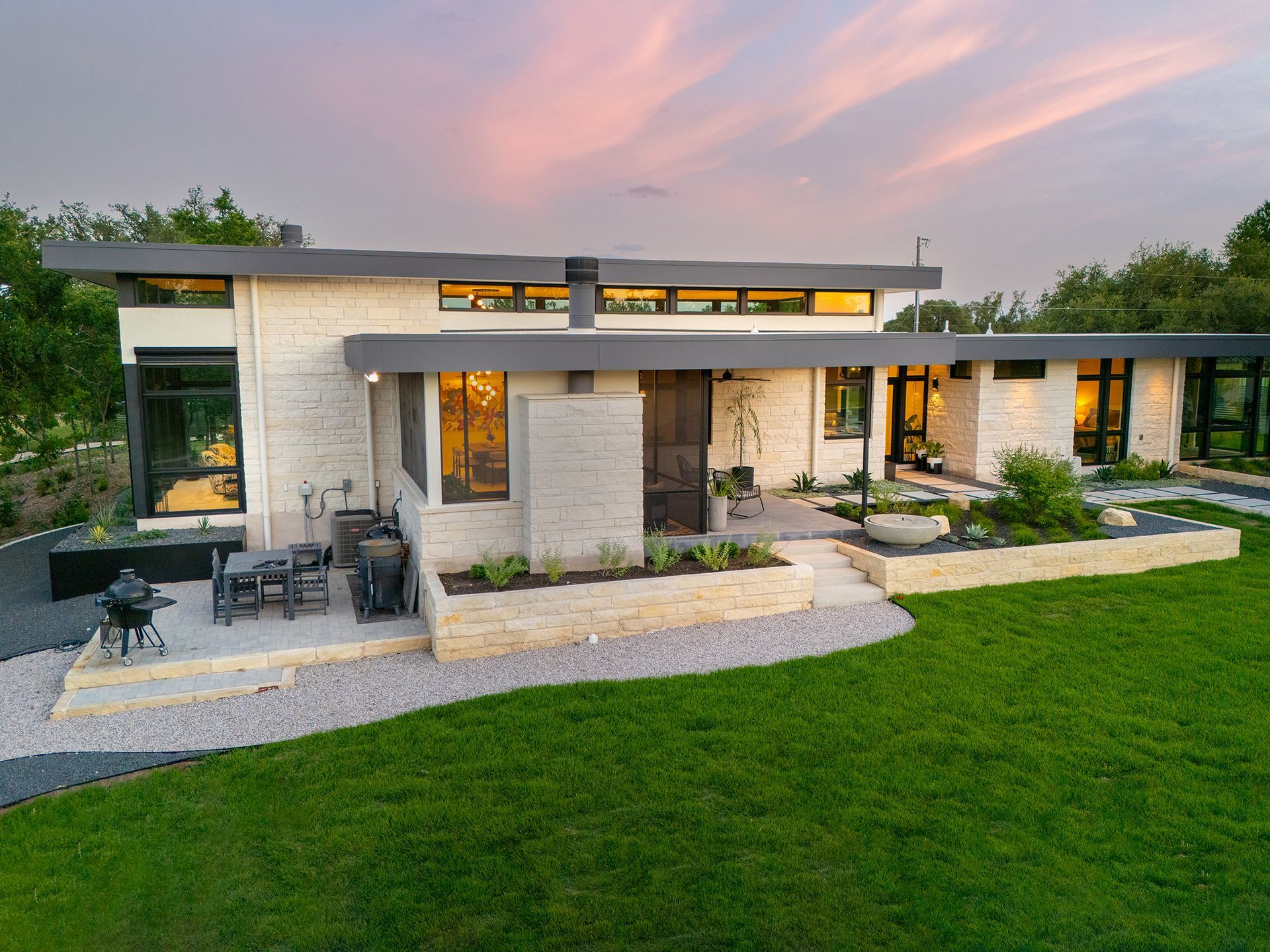 Modern home with light brick exterior, large windows, patio, and green lawn at dusk.