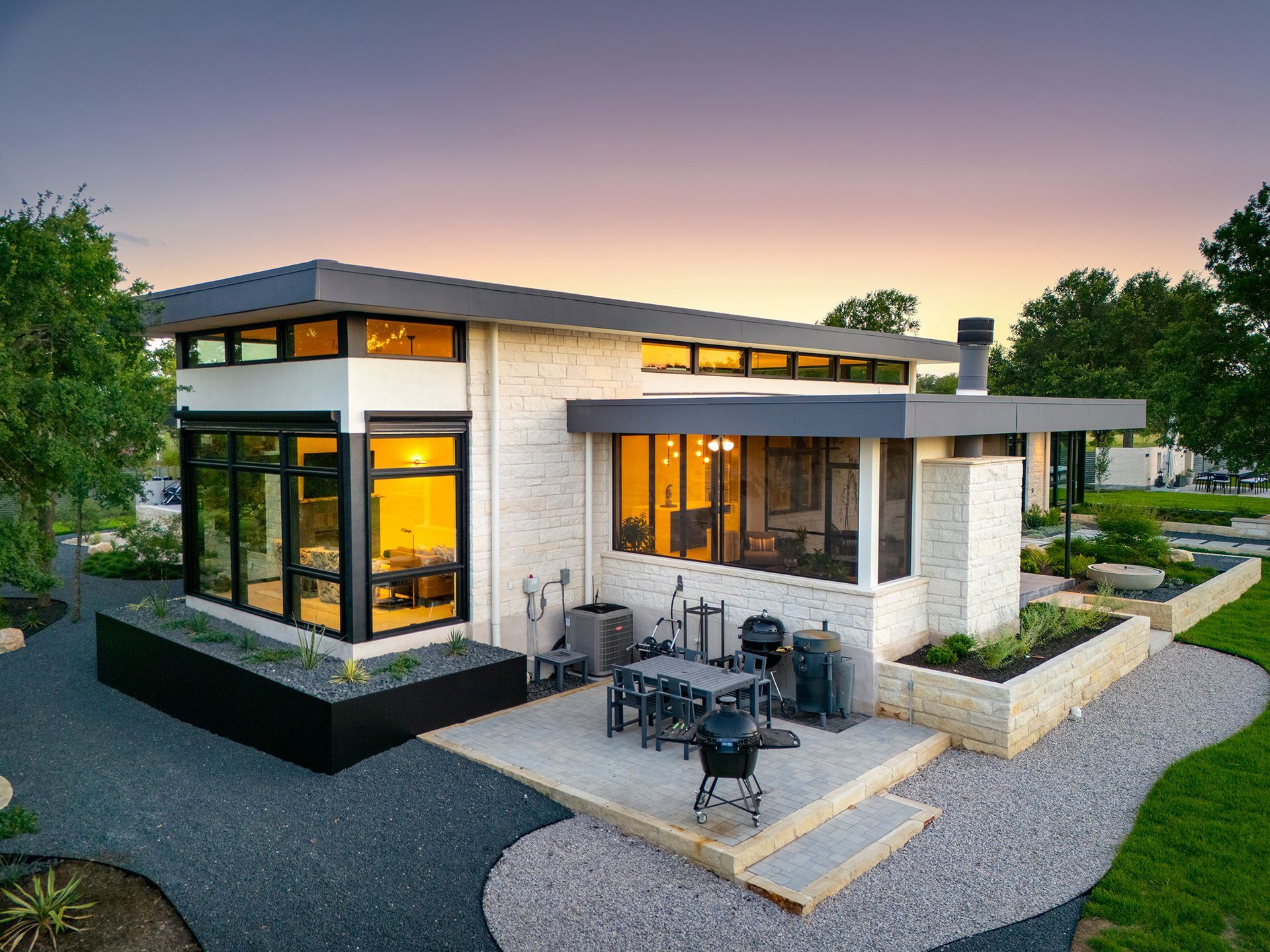 Modern, single-story house with large windows, patio, and landscaped yard at sunset.