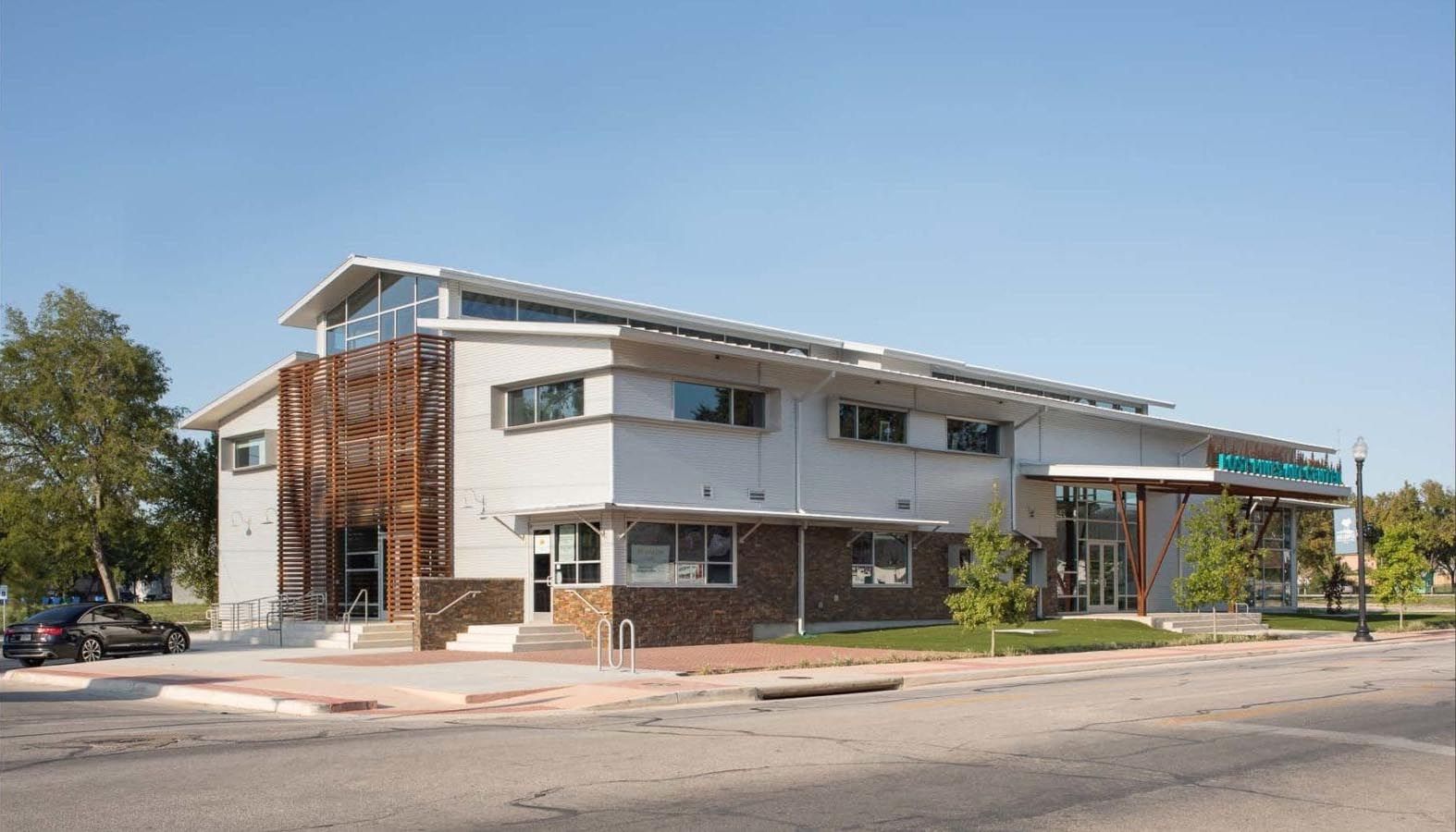 Modern two-story building with a light-colored exterior and a wooden facade section. Green signage at entrance, blue sky.