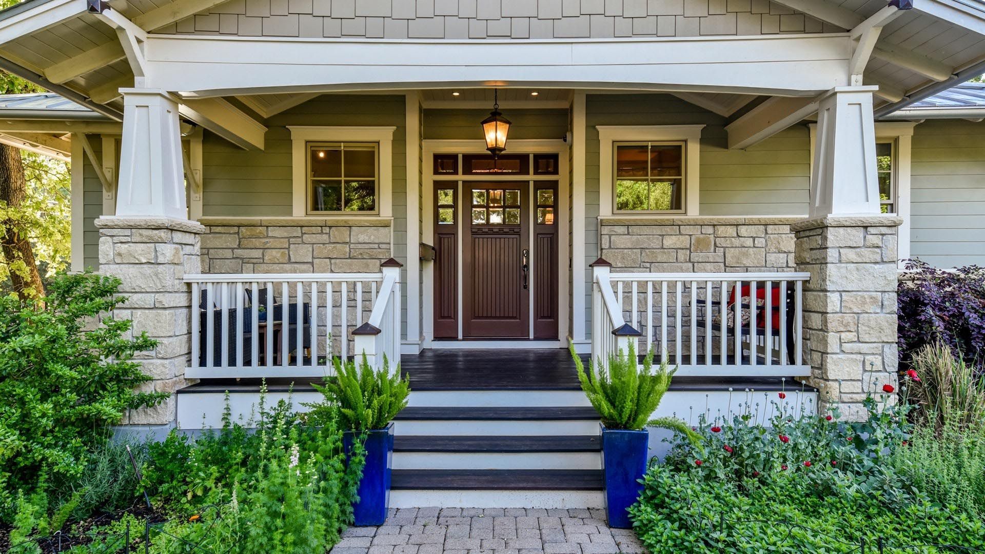 Green house with covered porch, stone accents, brown door, and blue planters.