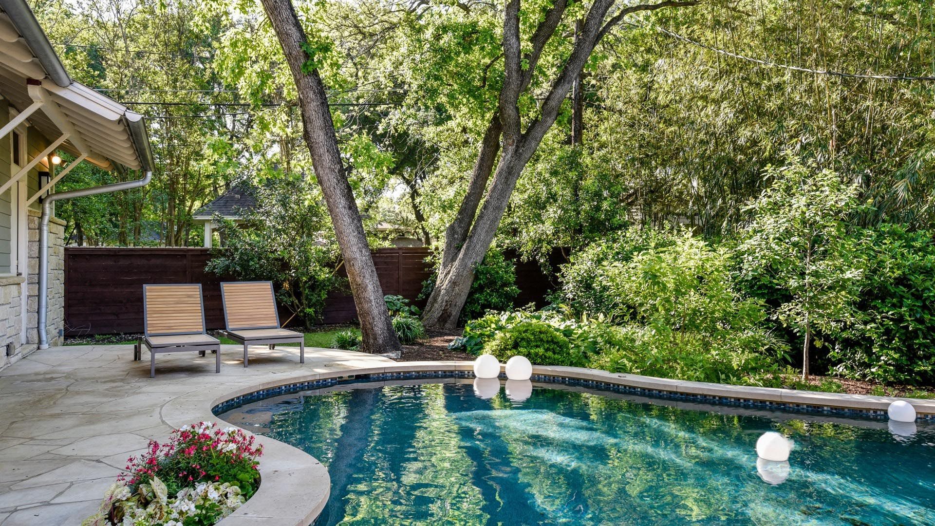 Backyard with pool, lounge chairs, lush greenery, and trees.