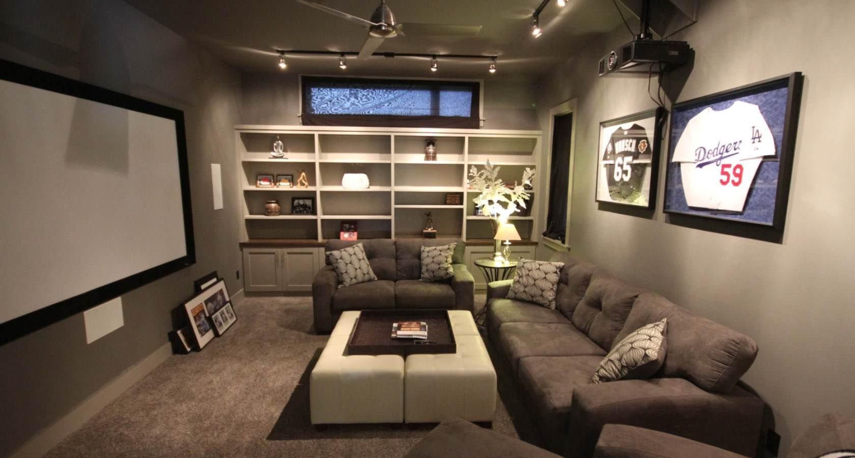 Cozy home theater with gray walls, sectional sofa, built-in shelves, projector screen, and framed sports jerseys.