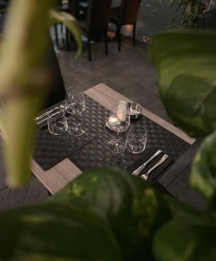 Apparecchiatura della tavola: bicchieri, posate, candela e tovagliette intrecciate. Il fogliame verde incornicia la scena.