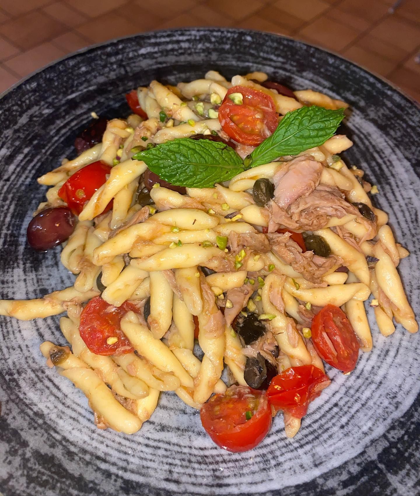 Piatto di pasta con pomodorini, olive, capperi e tonno, guarnito con menta su un piatto scuro.
