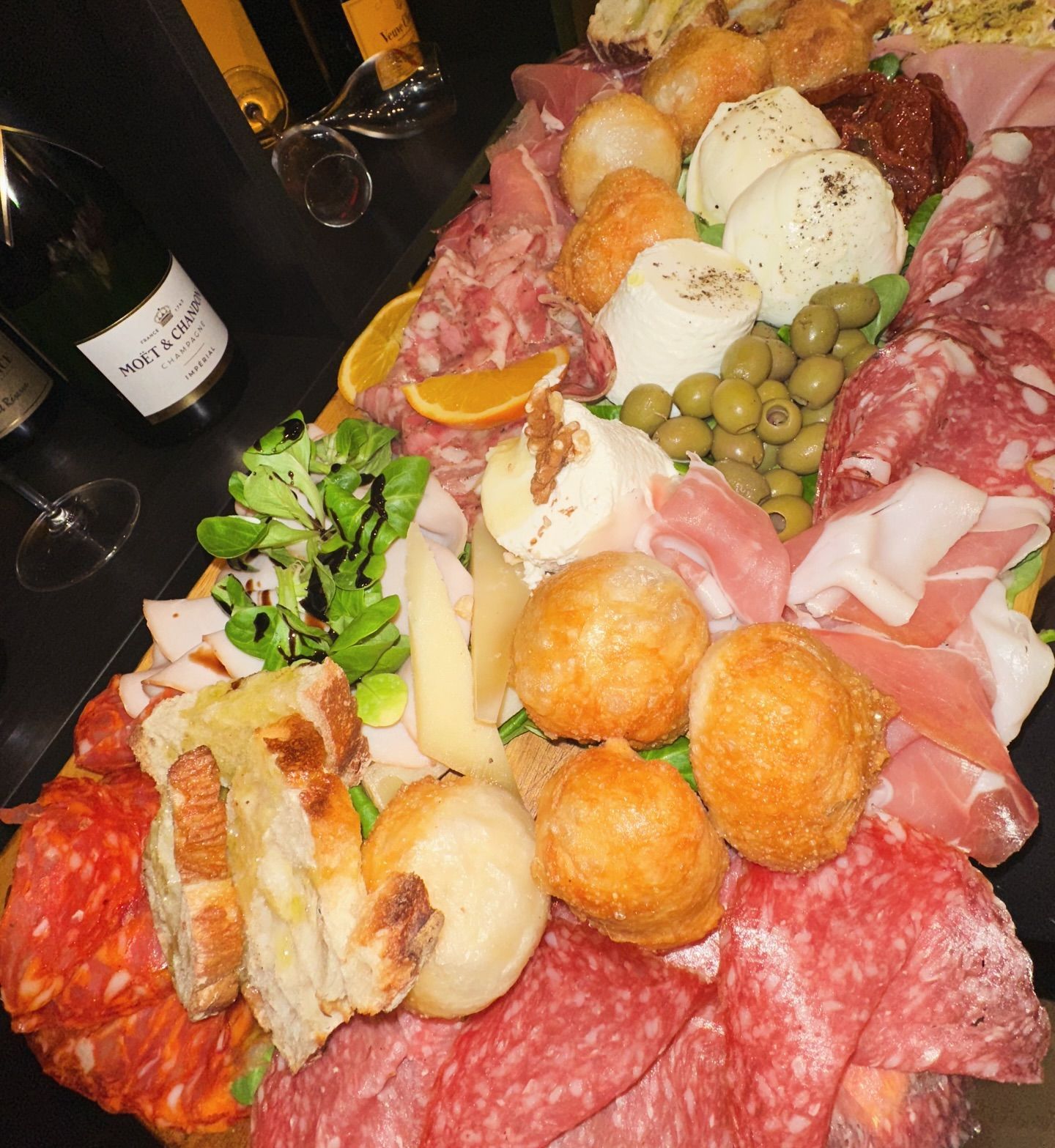 Tagliere di salumi con carne, formaggi, olive, pane e verdure; una bottiglia di vino.