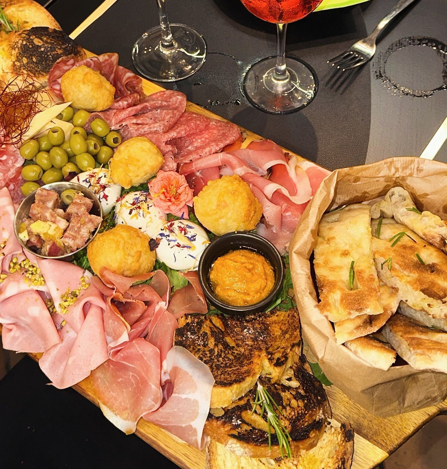 Tagliere di salumi con carni, formaggi, olive, pane e salsa, servito con bicchieri da vino.