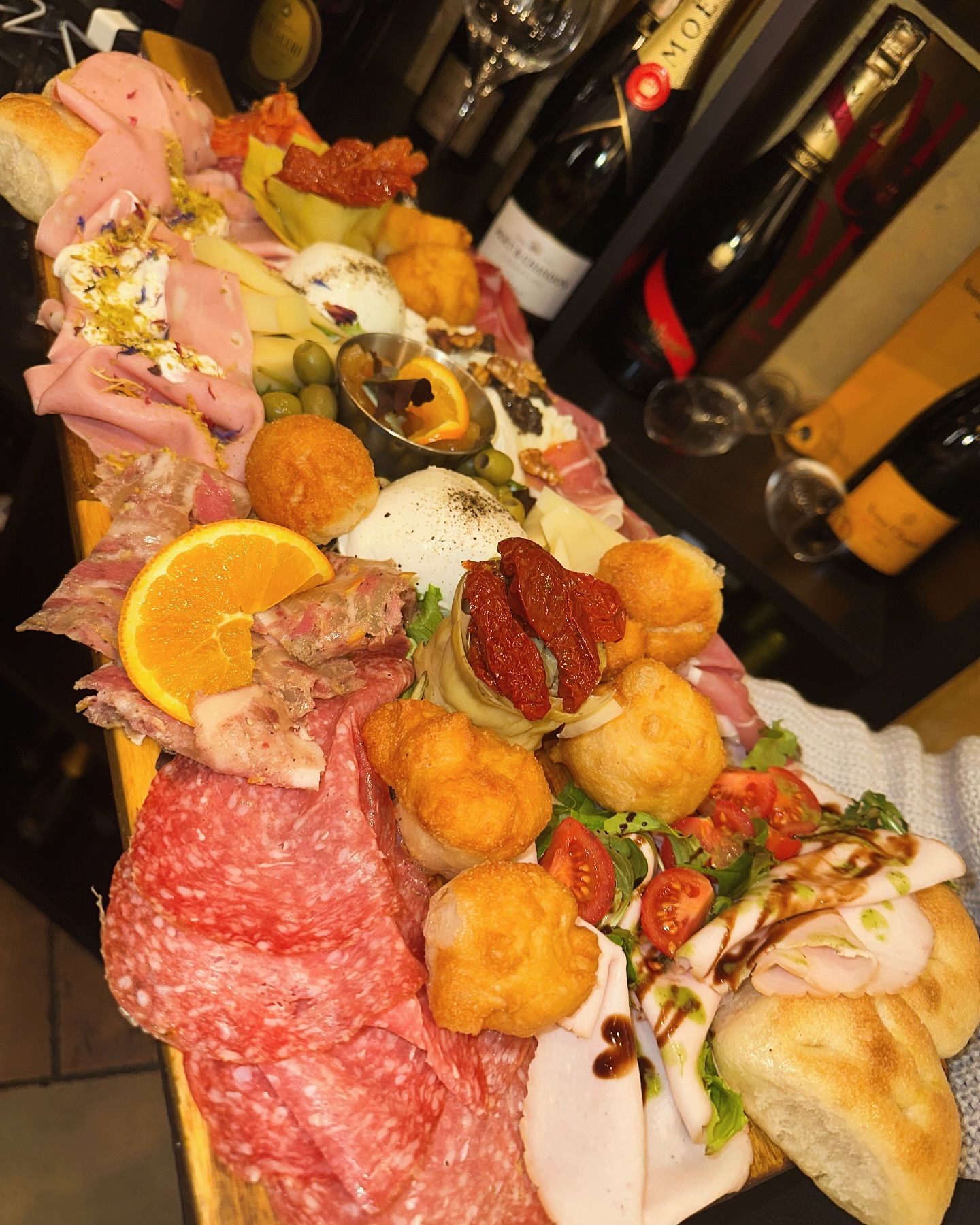 Tagliere di: salumi, formaggi, polpette fritte, pomodori secchi, olive, pane. Bottiglie di vino sullo sfondo.
