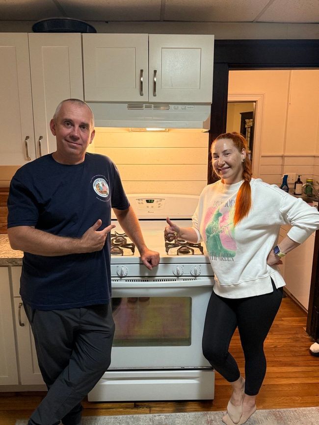 A man and woman stand by a white stove in a kitchen. They smile, pointing at the appliance.