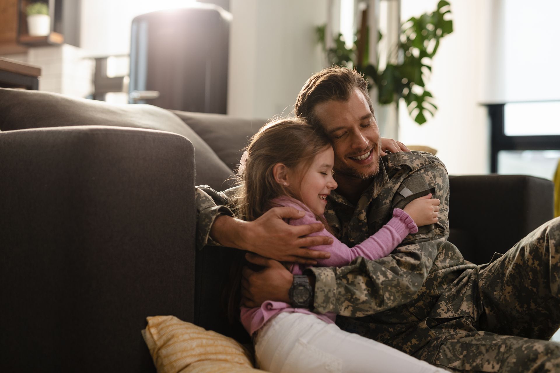 Man in camouflage embraces a young girl on a living room floor; both smiling.