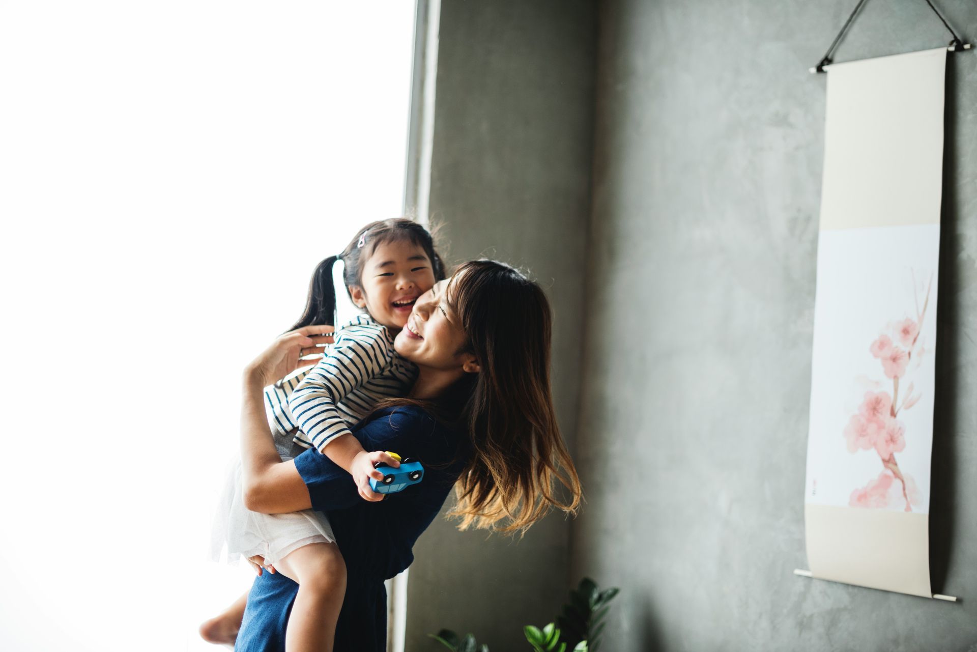 Woman holding a child on her back, both smiling. They're near a window, with a painting on the wall.