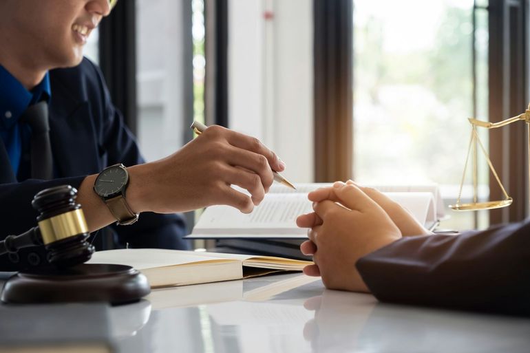 Lawyer points to open book, consulting with a client; gavel and scales of justice on the table.