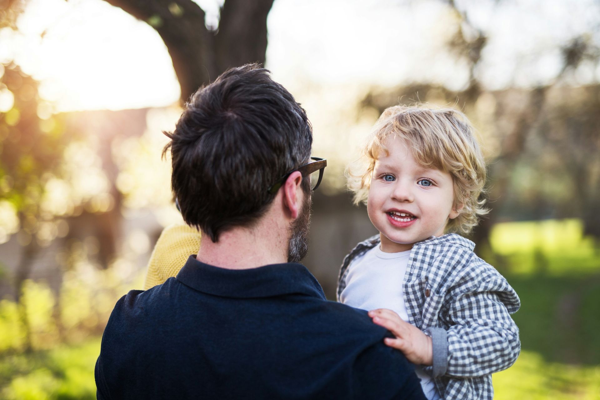 A person holding a child outdoors; the child smiles at the camera, sunlight in the background.