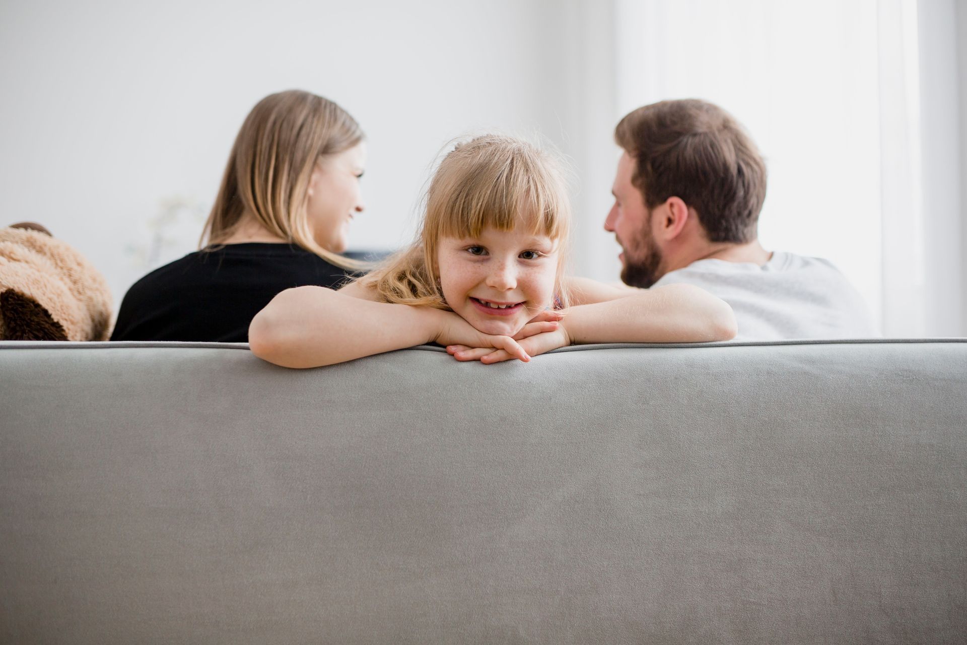 Girl smiling, leaning on a gray couch with parents in background. Bright room.