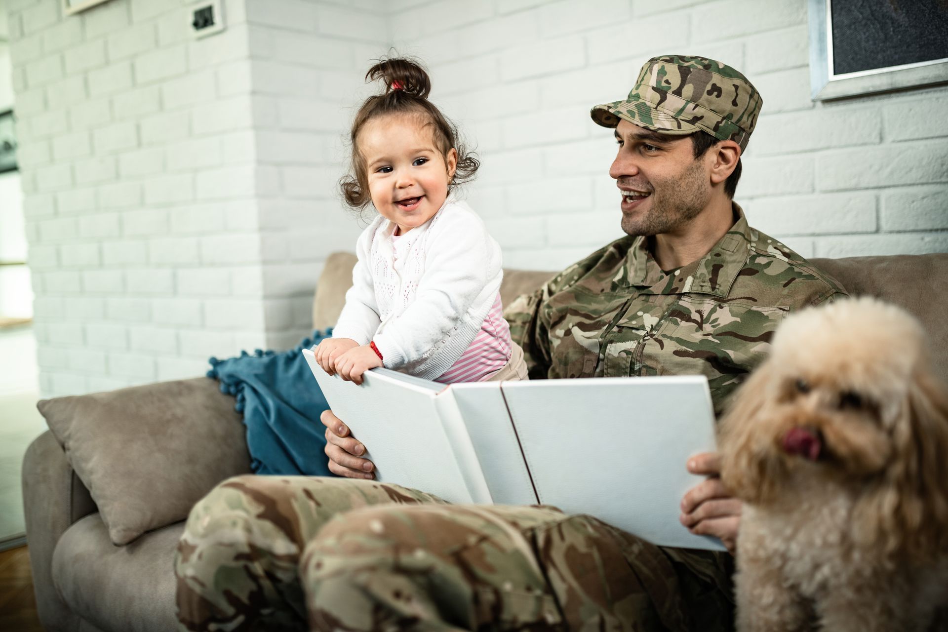 Man in camouflage reads to a toddler on a couch, with a dog.