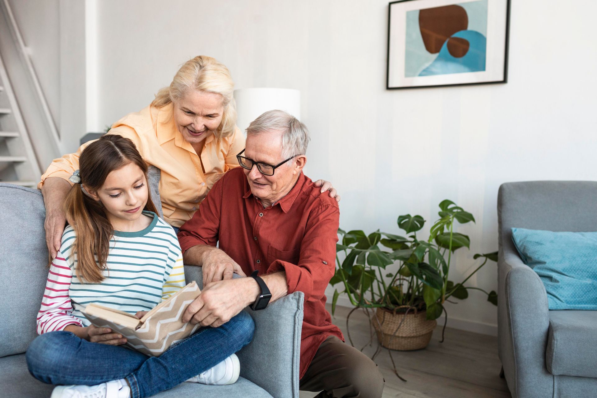 Girl reading book with grandparents on a couch; indoor setting.