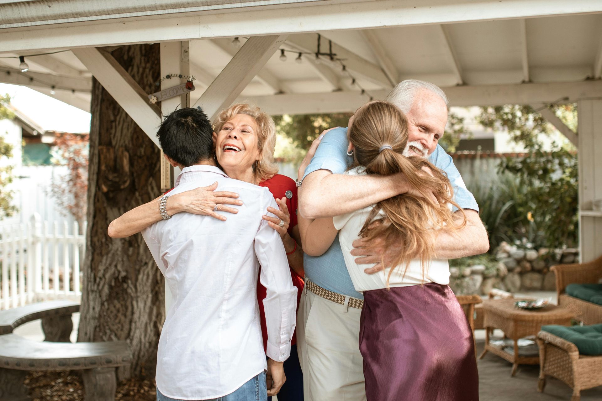 Two couples embrace outdoors, on a porch. Smiles and happy expressions, sunny day.