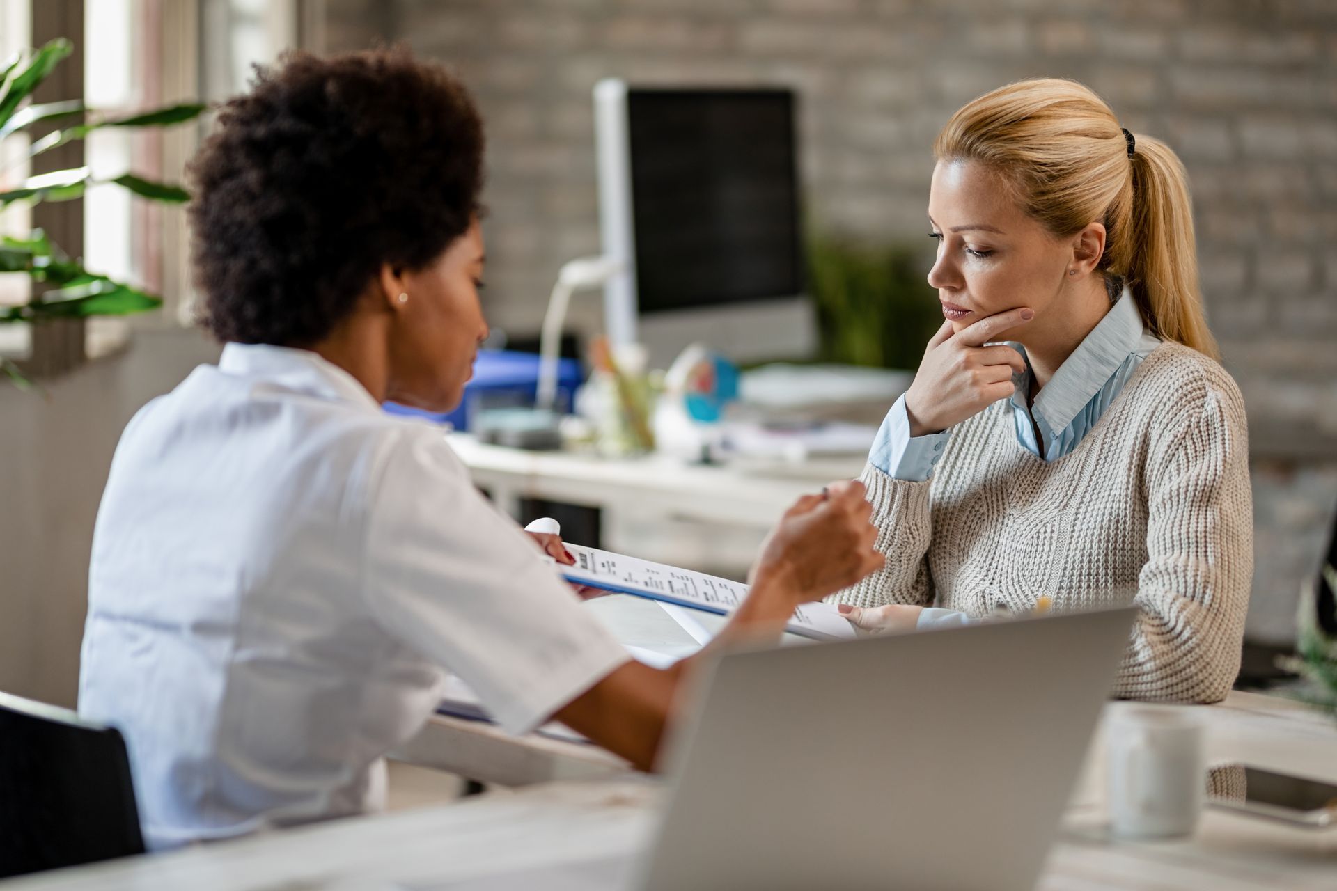 Woman consulting with medical professional; documents and laptop on desk.