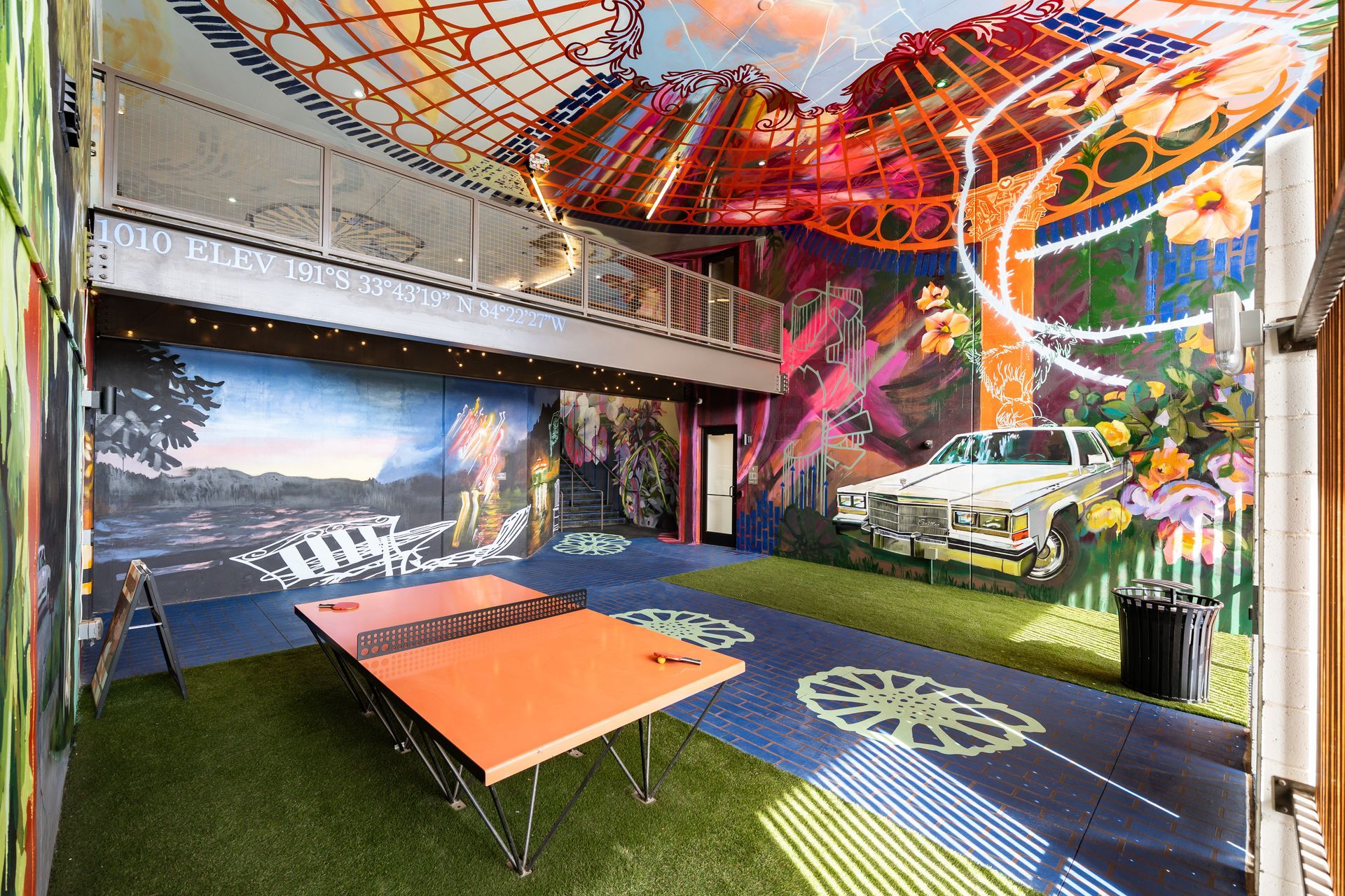 An indoor game room featuring an orange ping-pong table on artificial turf, surrounded by colorful muraled walls and floor.