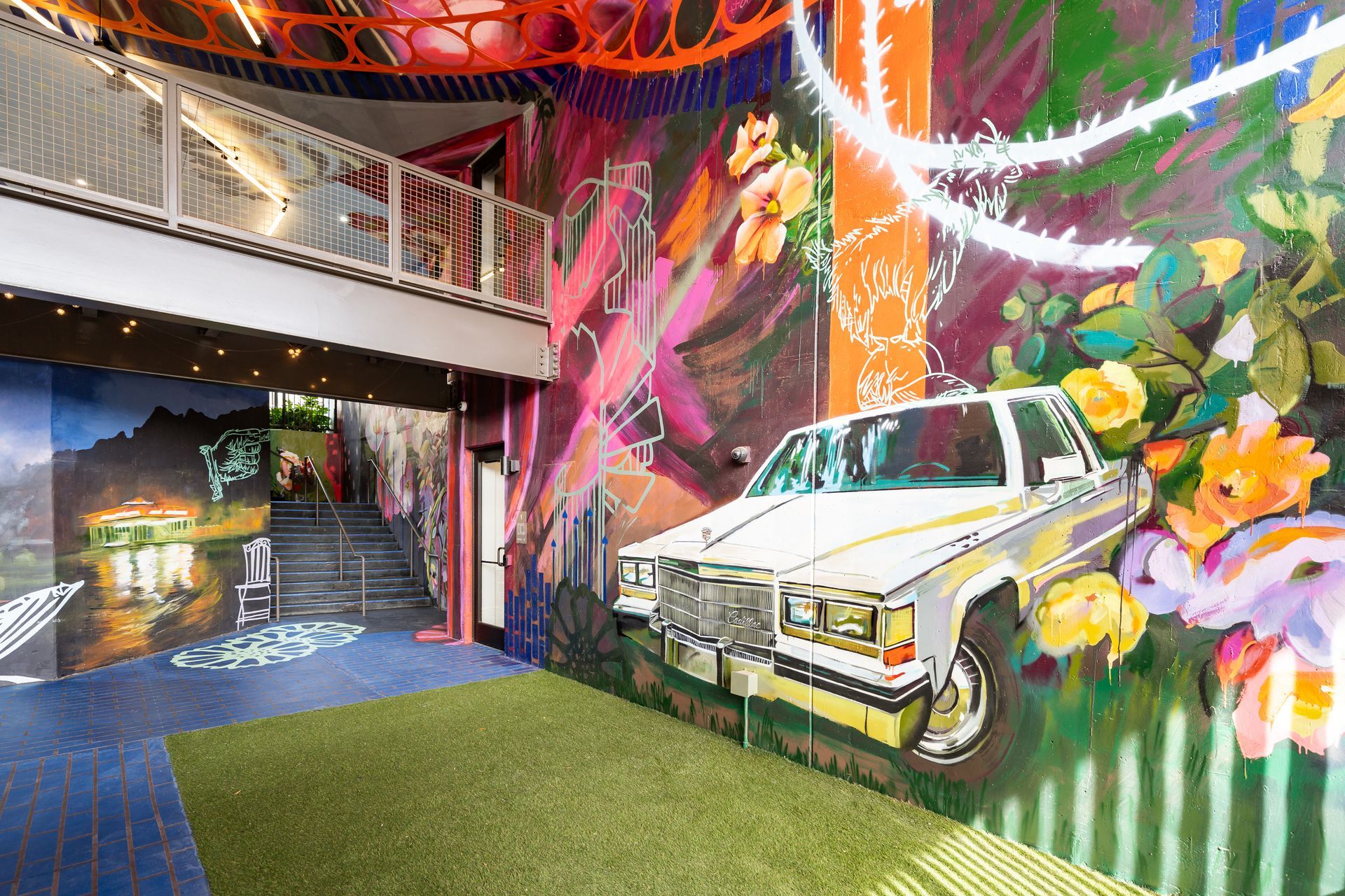 An indoor space features a wall mural of a white car, vibrant flowers, and an area covered with artificial green grass.