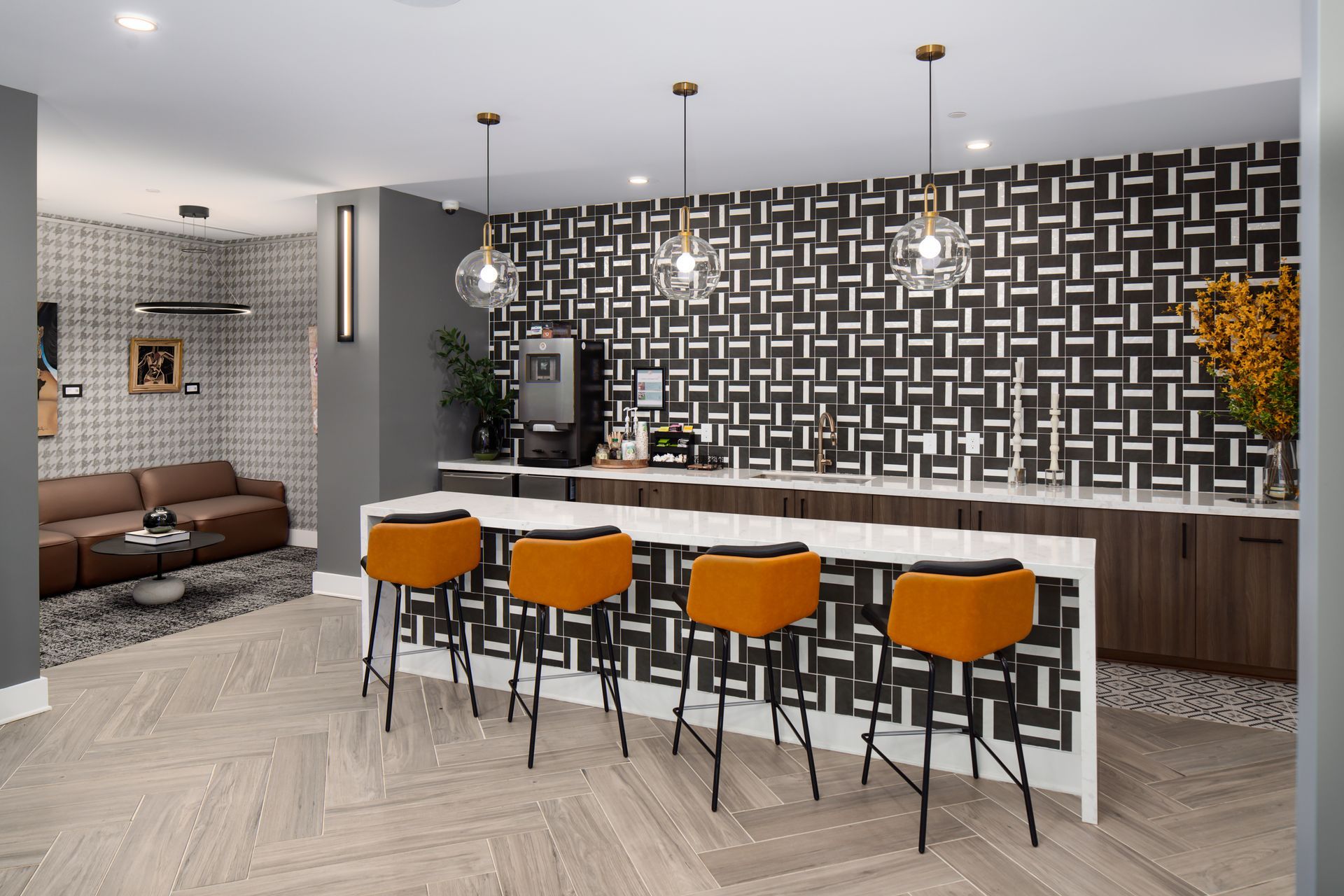 Modern lounge area with a coffee station, white bar island, four orange stools, and geometric black-and-white wallpaper.