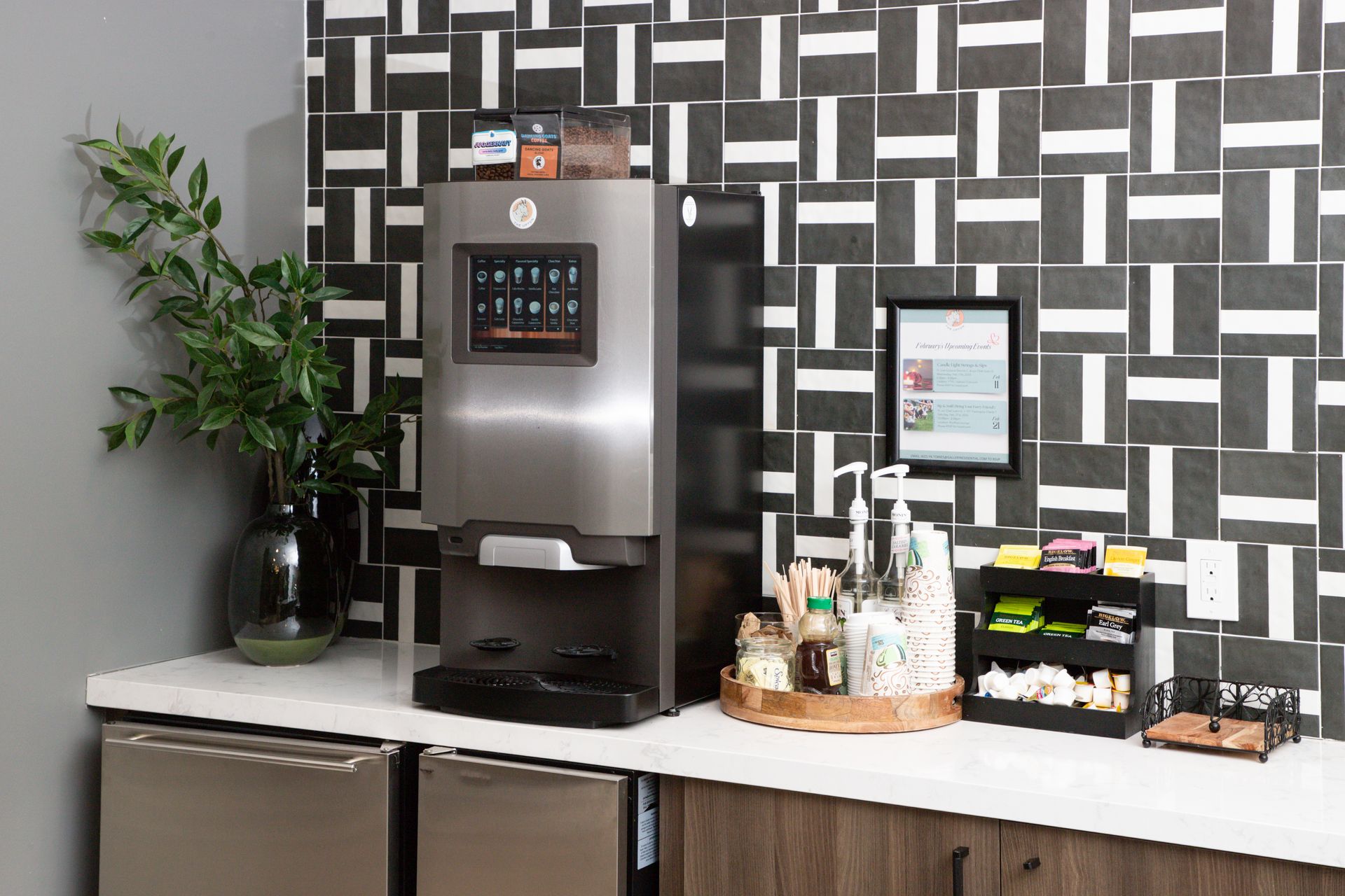 A stainless steel coffee machine sits on a white counter next to a plant, condiments, and a small snack shelf.
