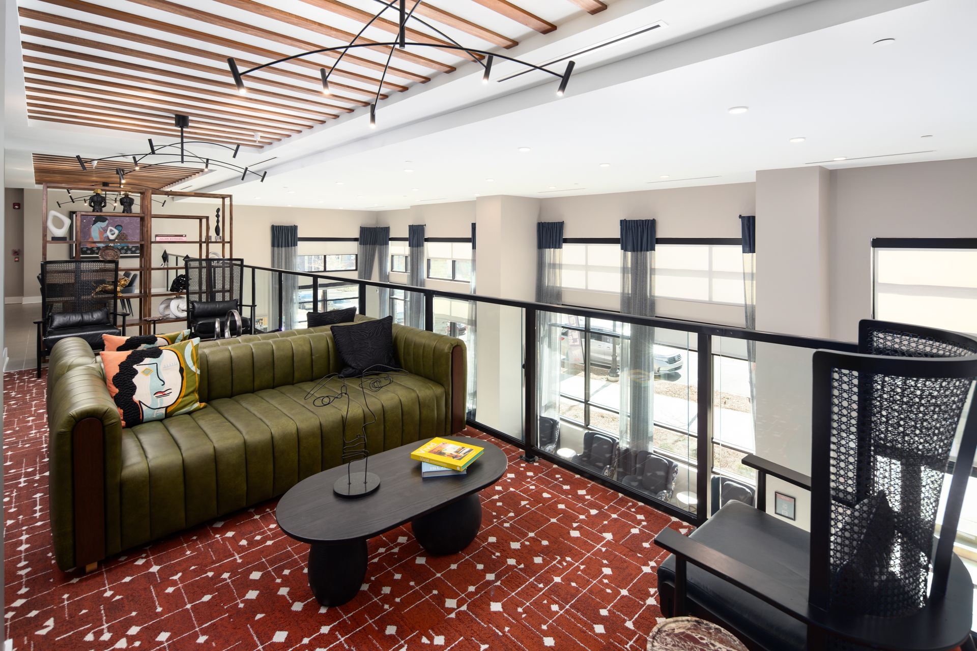 Modern lounge area with a green sofa, oval coffee table, and patterned red carpet, overlooking a bright indoor space.