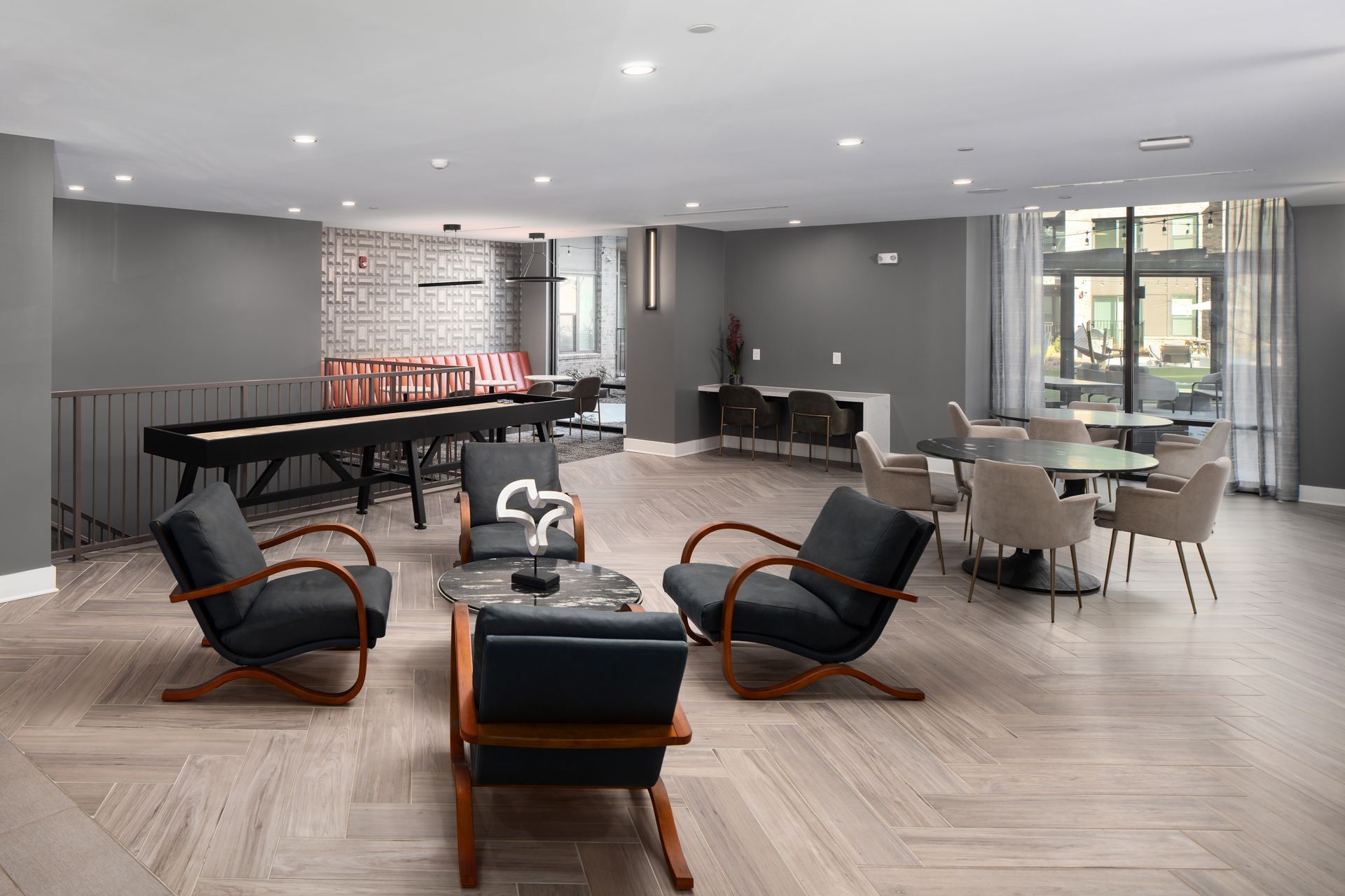 A modern lounge area with dark armchairs, a shuffleboard table, a small dining set, and light herringbone wood floors.