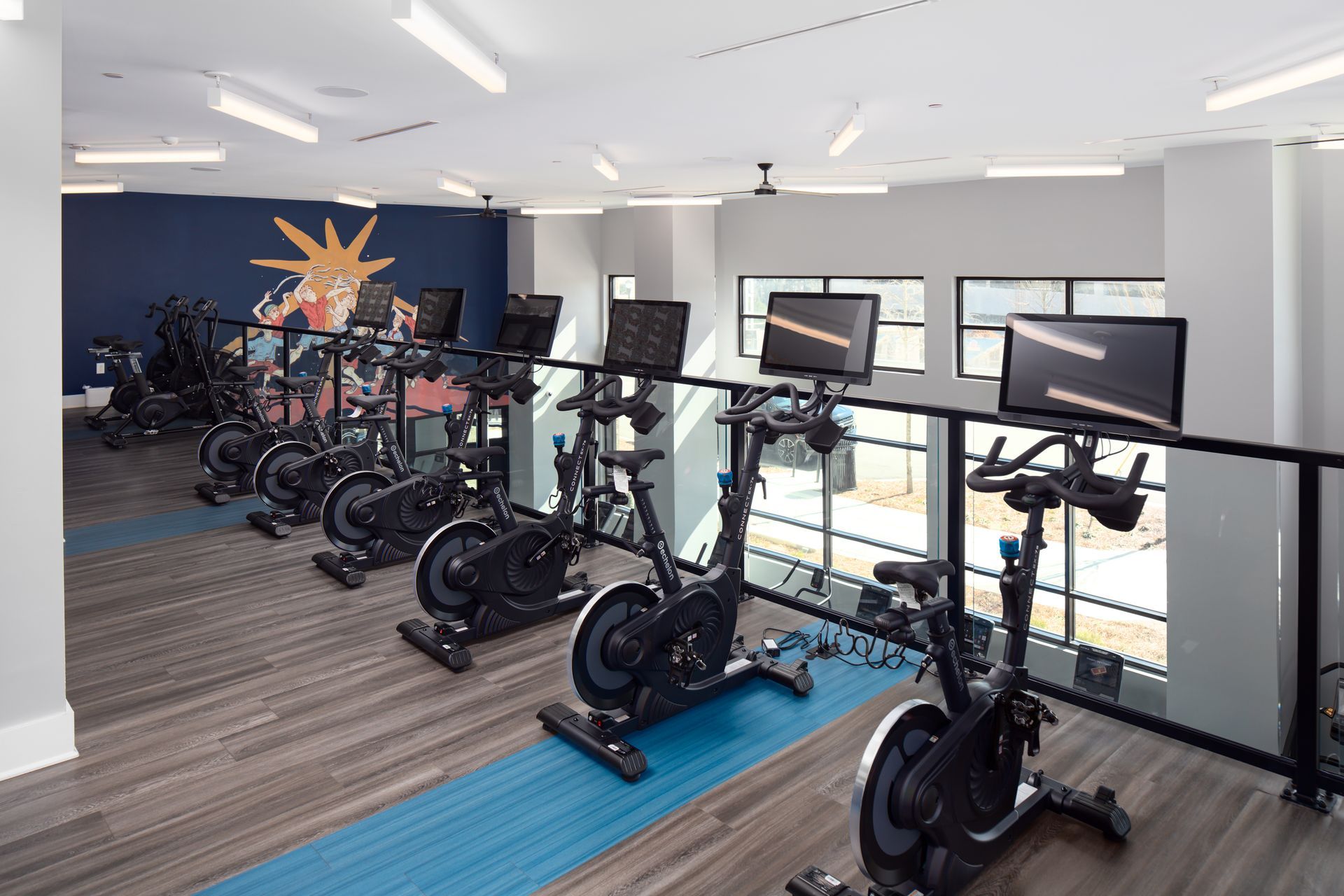 A row of stationary exercise bikes with monitors, arranged on a blue mat inside a fitness studio with a wall mural.