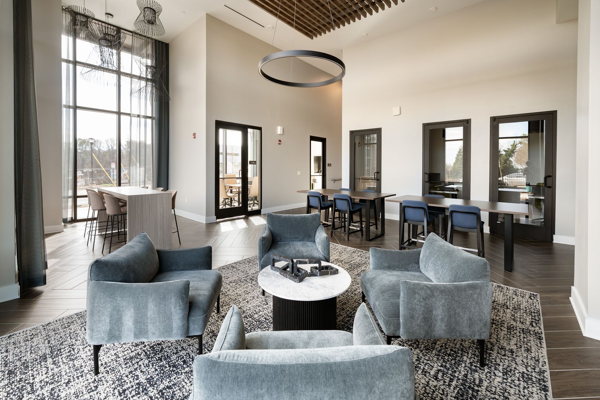 Modern lobby with blue armchairs, a round marble coffee table, wooden dining tables, and large windows with sheer curtains.