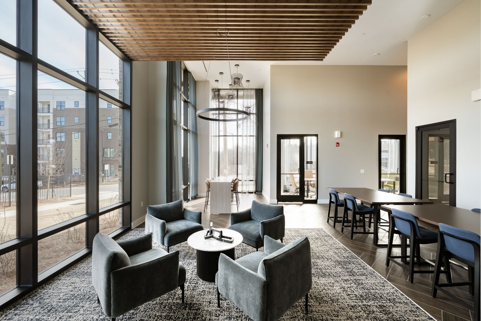 Modern, sunlit lounge with gray armchairs around a circular table, near wooden-slatted ceilings and large windows.