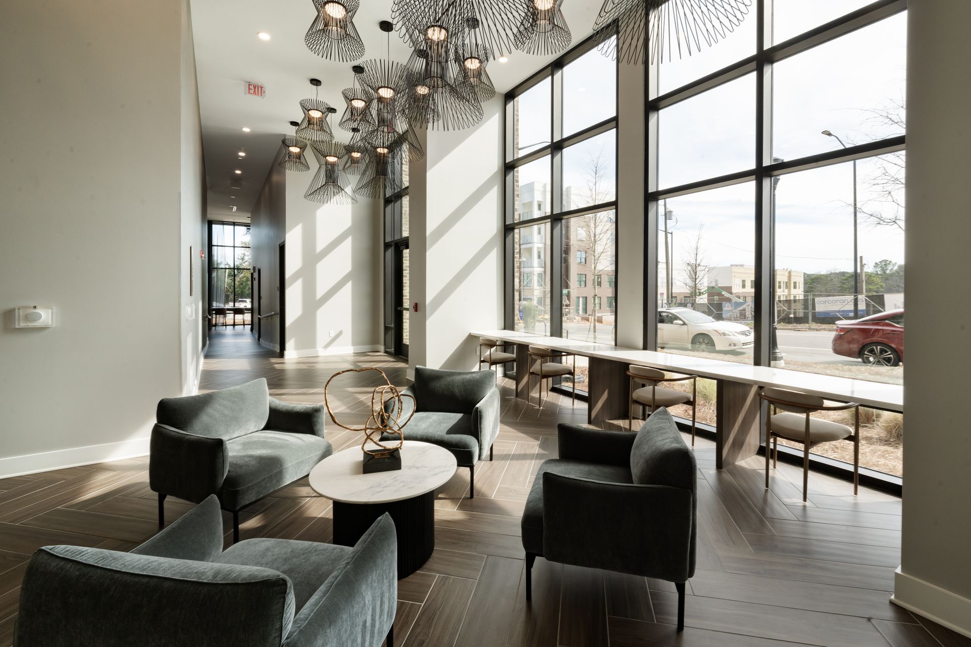 Modern lobby with lounge chairs, a central coffee table, and a long window-side counter under ornate light fixtures.