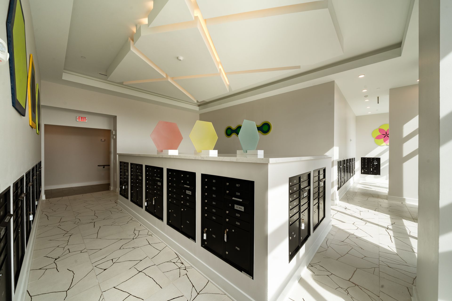 A modern mailroom with rows of black mailboxes, light-colored tiled flooring, and hexagonal wall art in a bright hall.