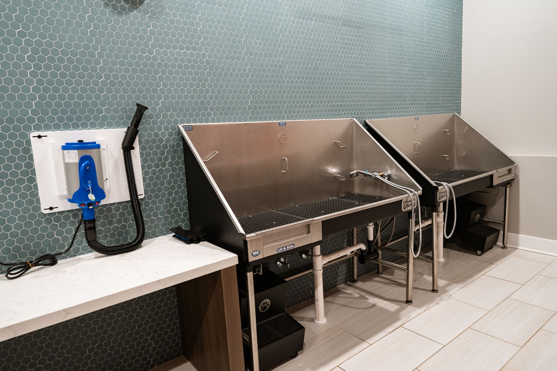 Two stainless steel pet washing stations with black rubber mats and a wall-mounted dryer in a room with teal wallpaper.