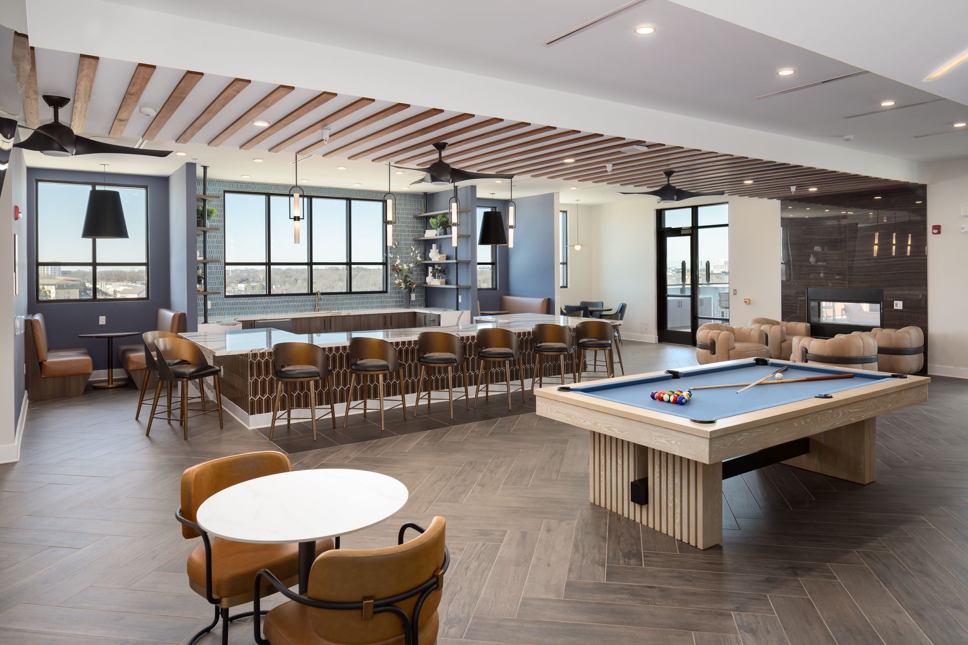 Modern lounge area with a pool table, bar seating, wood-slat ceiling, and circular tables on a herringbone wood floor.
