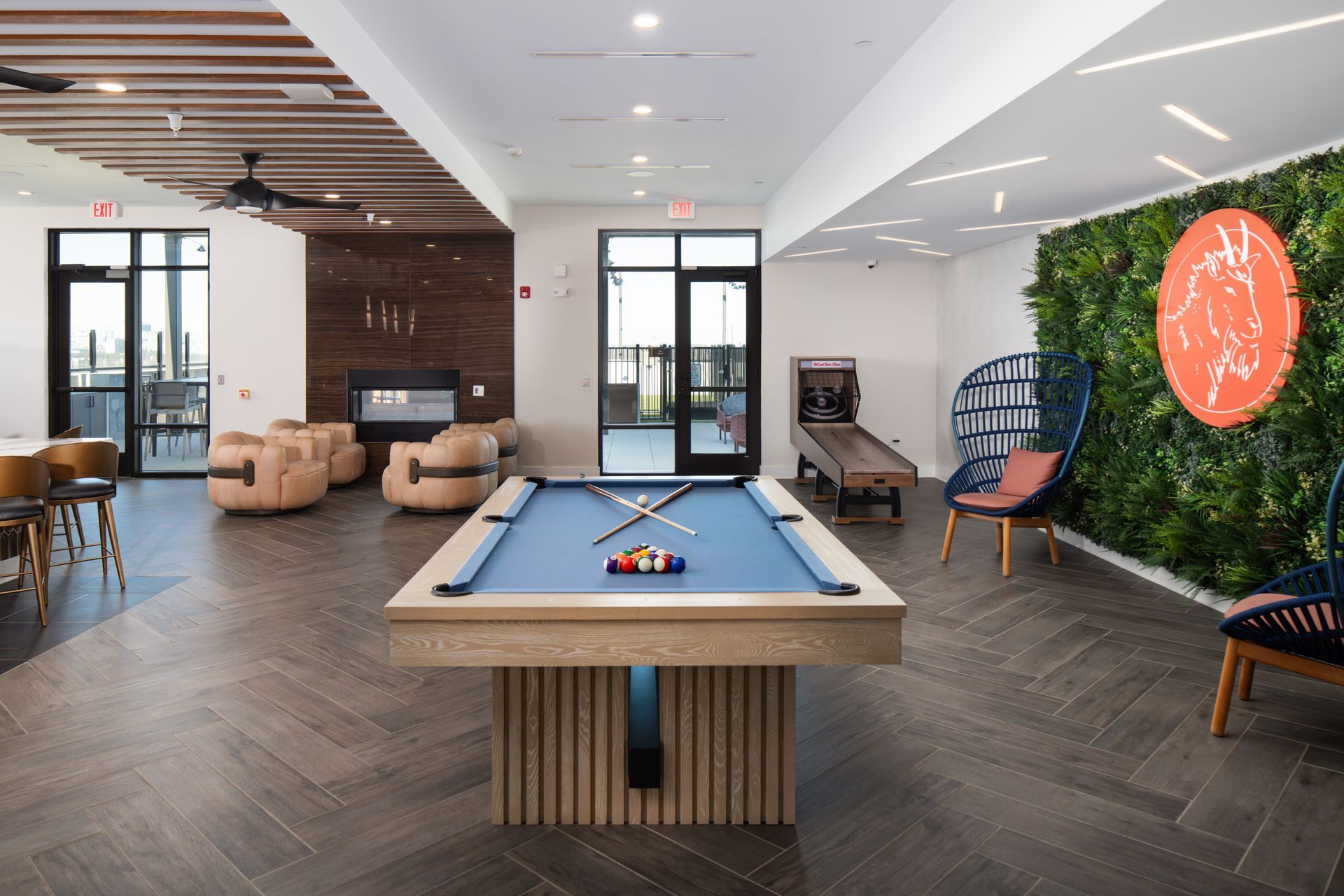 A modern game room with a blue pool table, lounge chairs near a fireplace, a skee-ball machine, and a green plant wall.