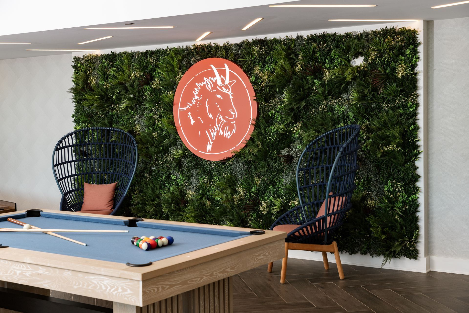 A pool table sits in front of a green plant wall featuring a round, orange sign with a white goat logo and two dark chairs.