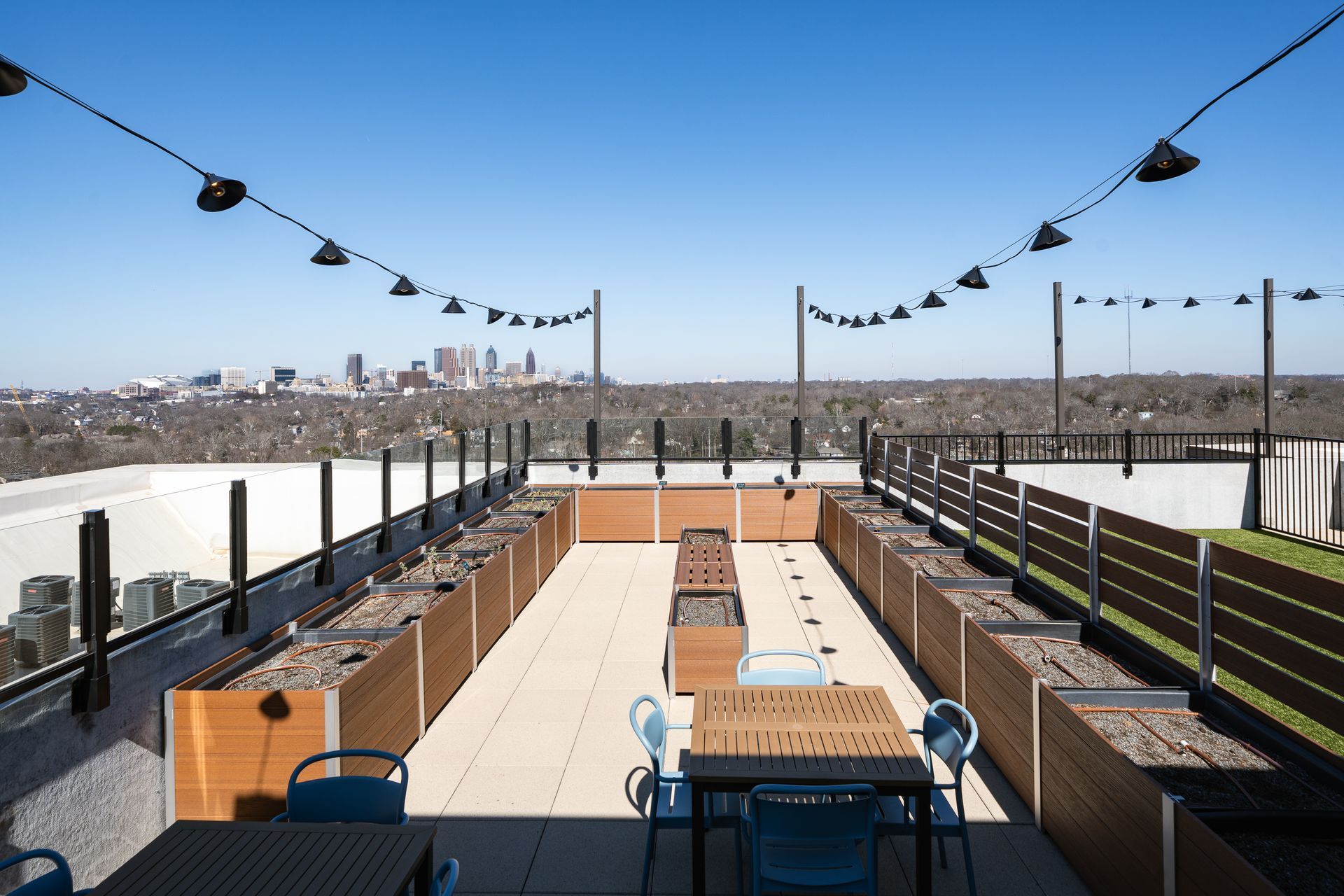 A rooftop terrace with wooden planters, tables, and blue chairs, featuring a clear view of a distant city skyline.
