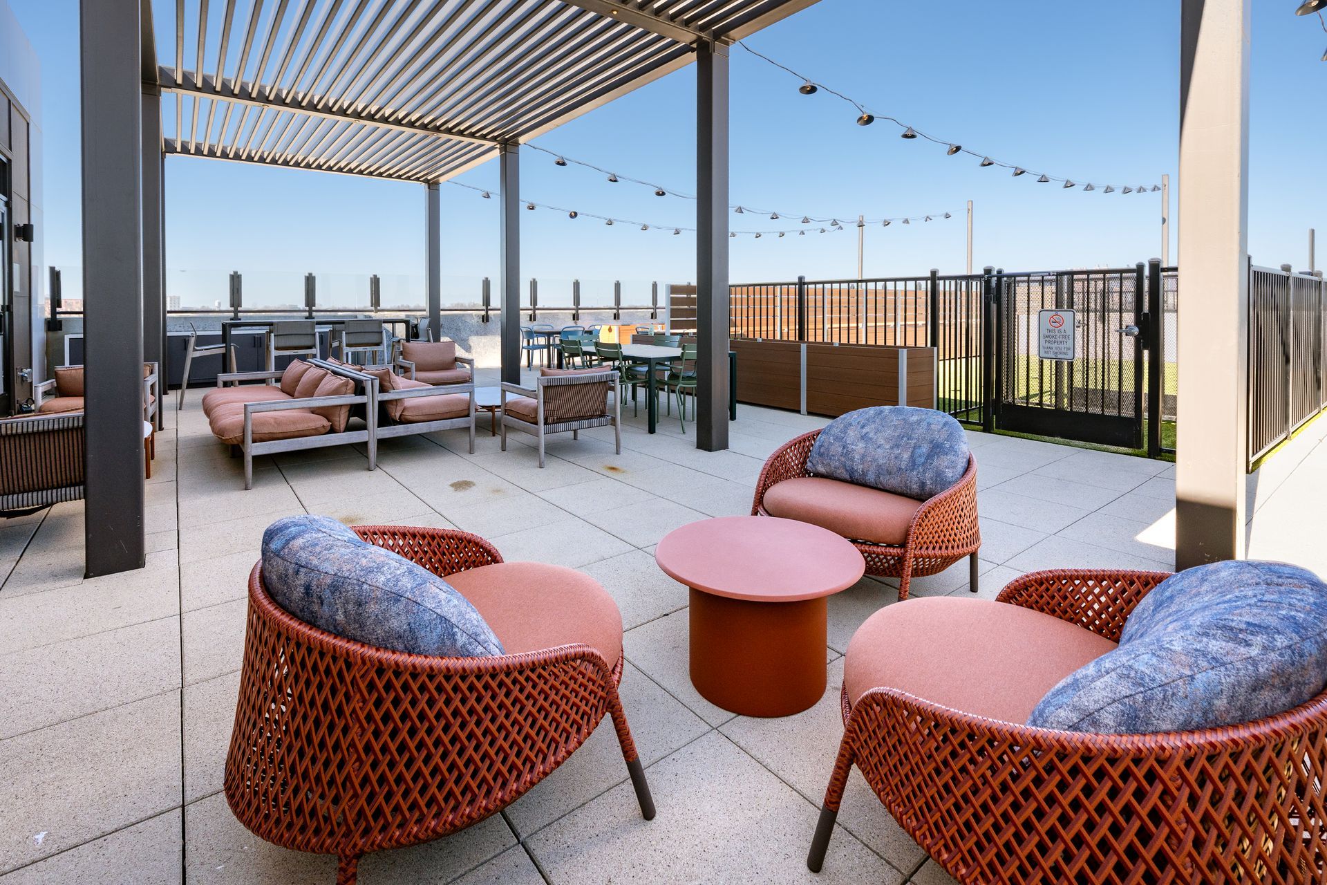 Outdoor rooftop terrace with woven armchairs, pink cushions, a matching circular coffee table, and a shaded pergola.
