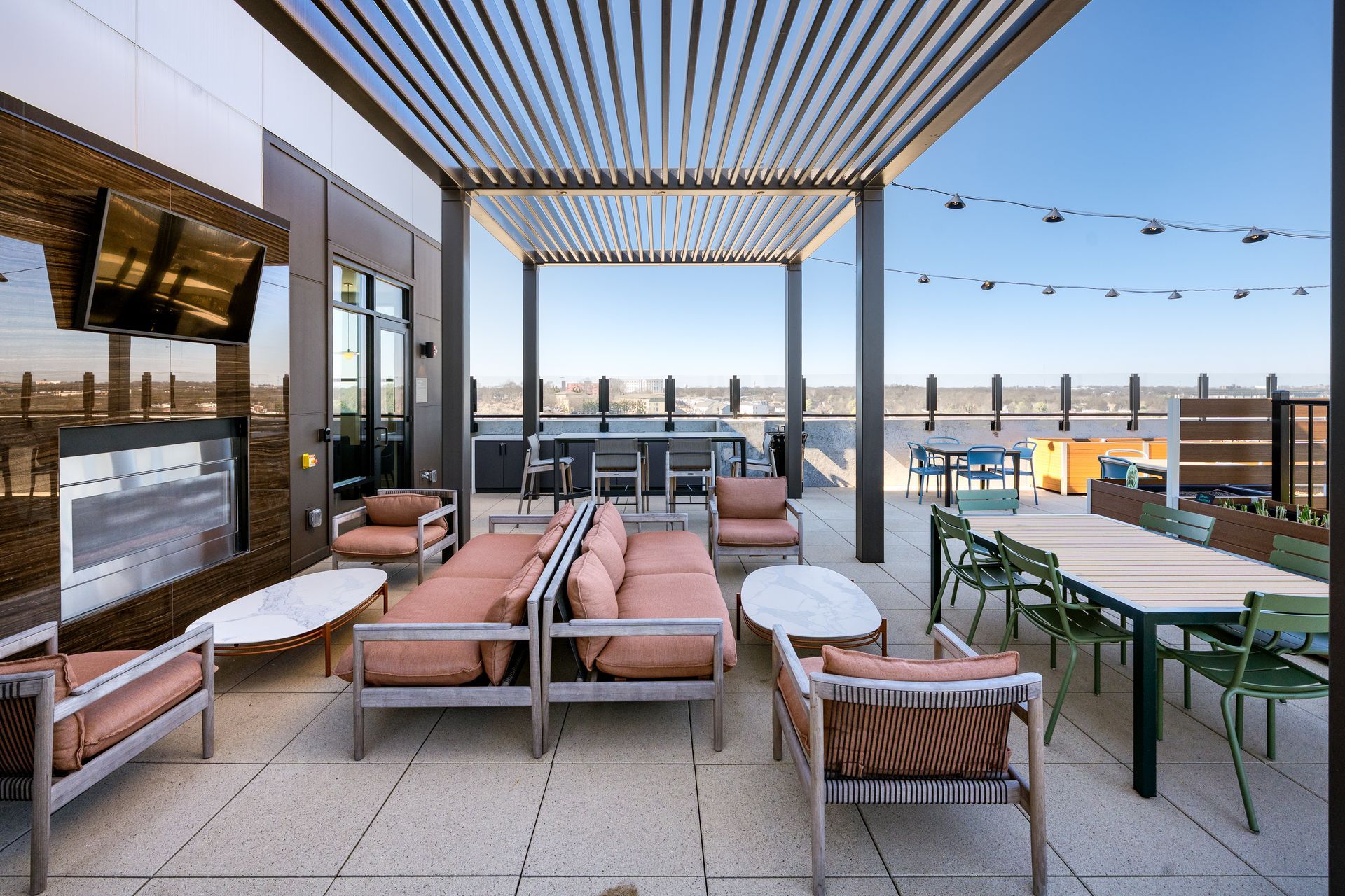 Modern rooftop patio with a fireplace, lounge seating, dining table, and pergola under a clear blue sky.