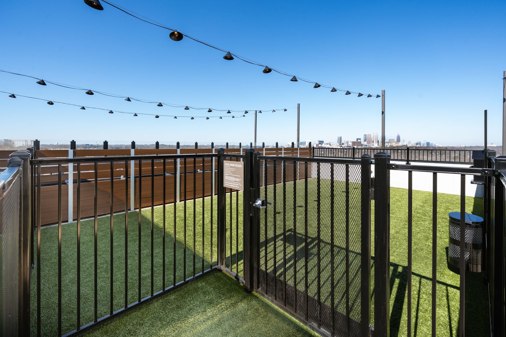 A gated rooftop dog run with artificial turf, surrounded by metal fencing under a clear blue sky with string lights.