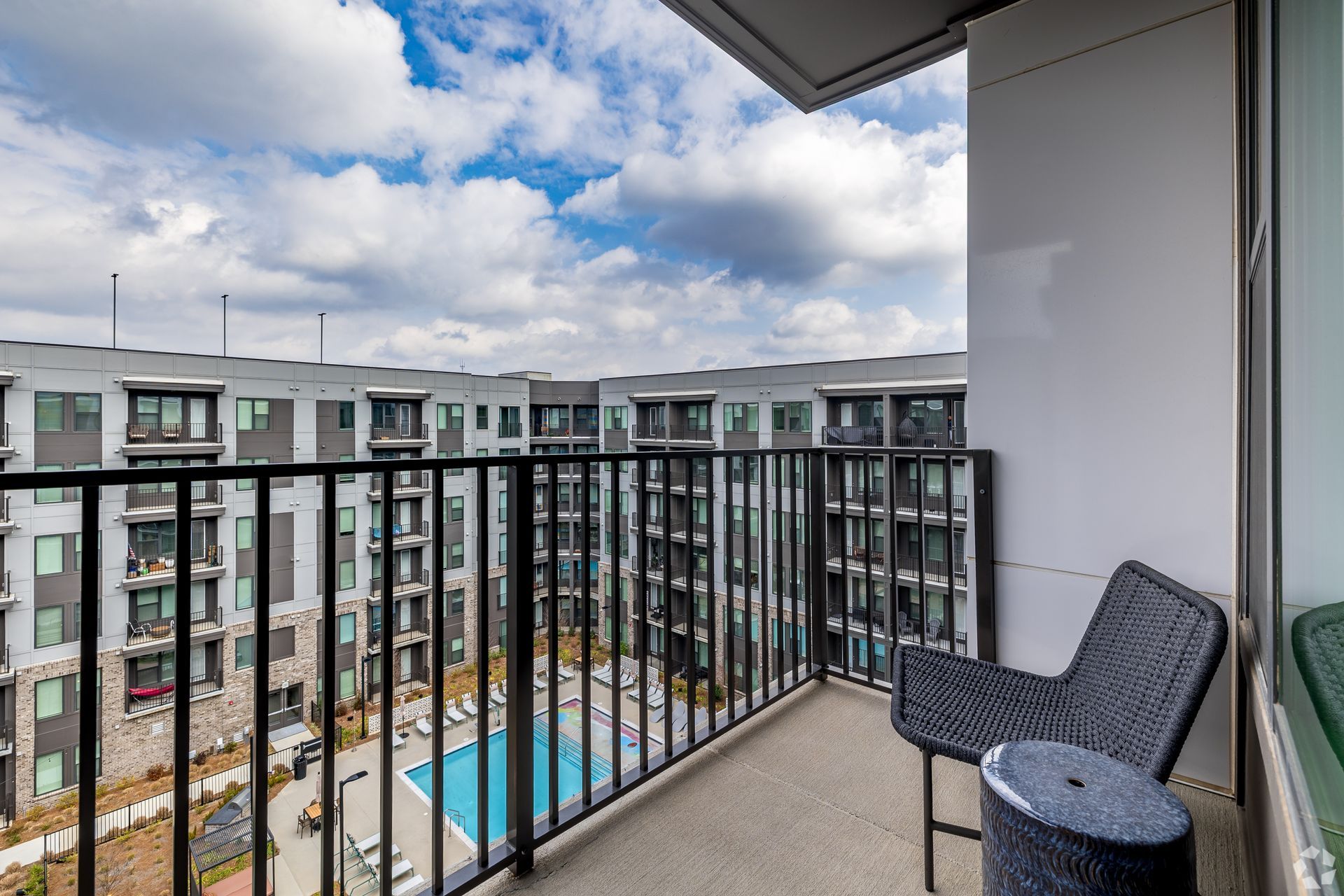 A balcony with a modern chair overlooks an apartment courtyard with a swimming pool under a partly cloudy sky.