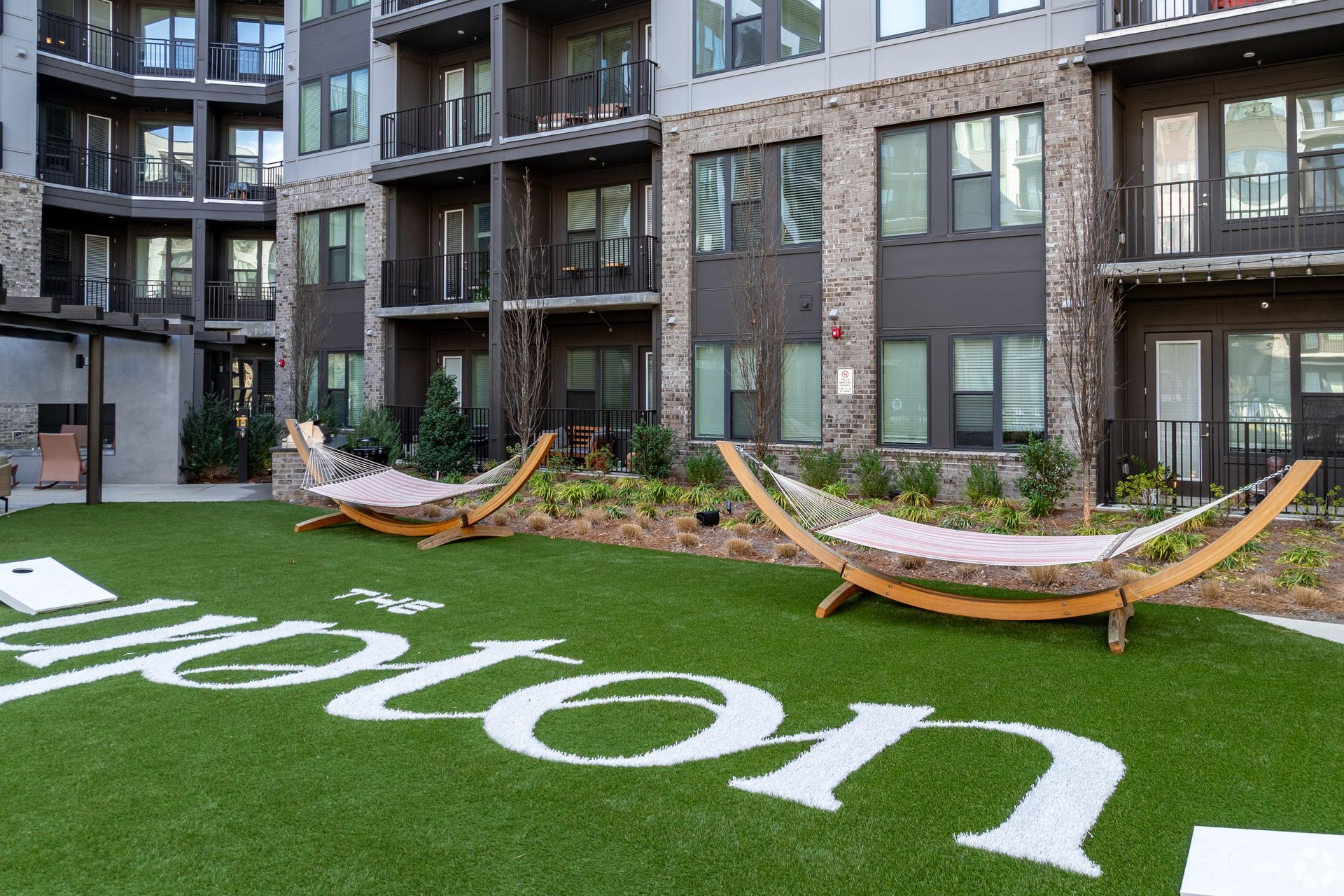 Two hammocks sit on a green lawn in front of a modern apartment building with a white logo painted on the grass.