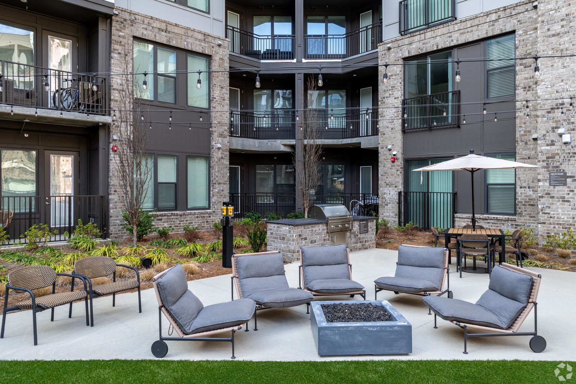 A courtyard patio with grey lounge chairs, a fire pit, and stone-accented apartment buildings in the background.