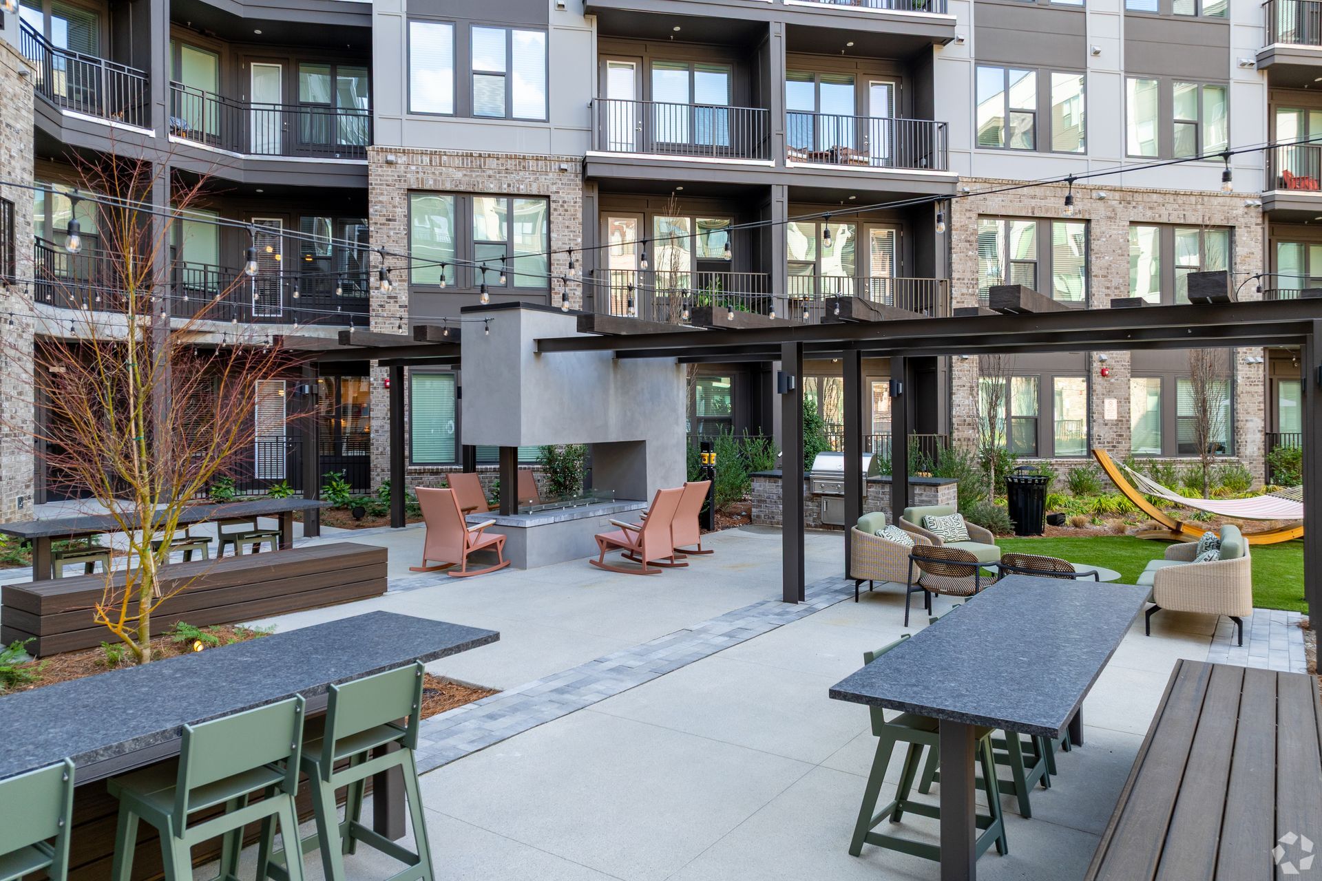 Outdoor apartment courtyard with a fireplace, lounge seating, dining tables, and a metal pergola under a multistory building.